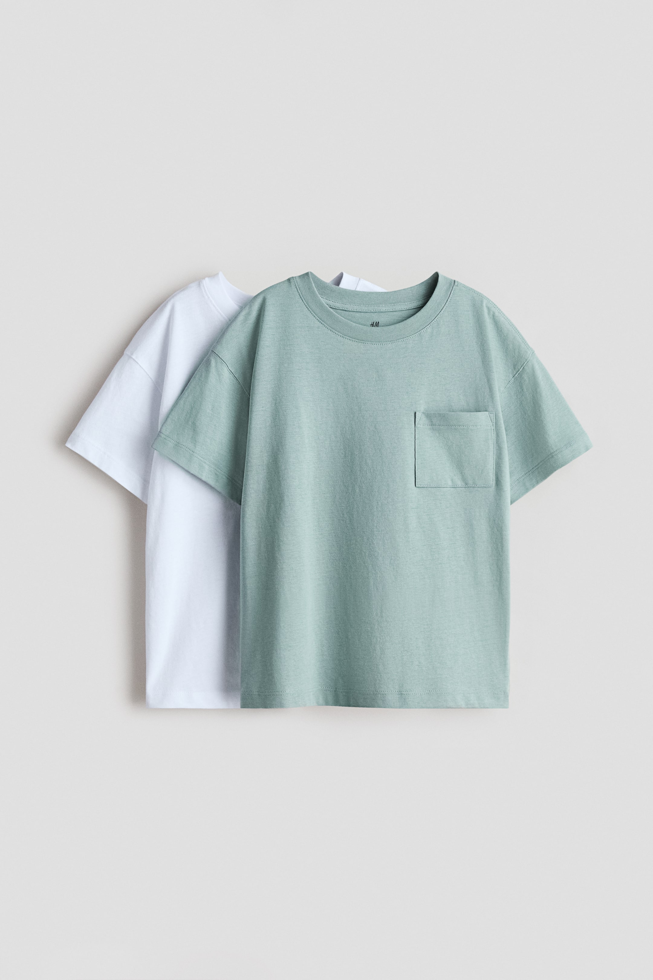 Vezi imaginea mai mare: Două tricouri supradimensionate sunt expuse, cu un tricou verde deschis prezentându-și partea din față peste un tricou alb. Ambele tricouri au umeri căzuți și o textură moale de jerseu din bumbac, în timp ce tricoul verde deschis din față are, de asemenea, un buzunar aplicat pe piept și un guler rotund.