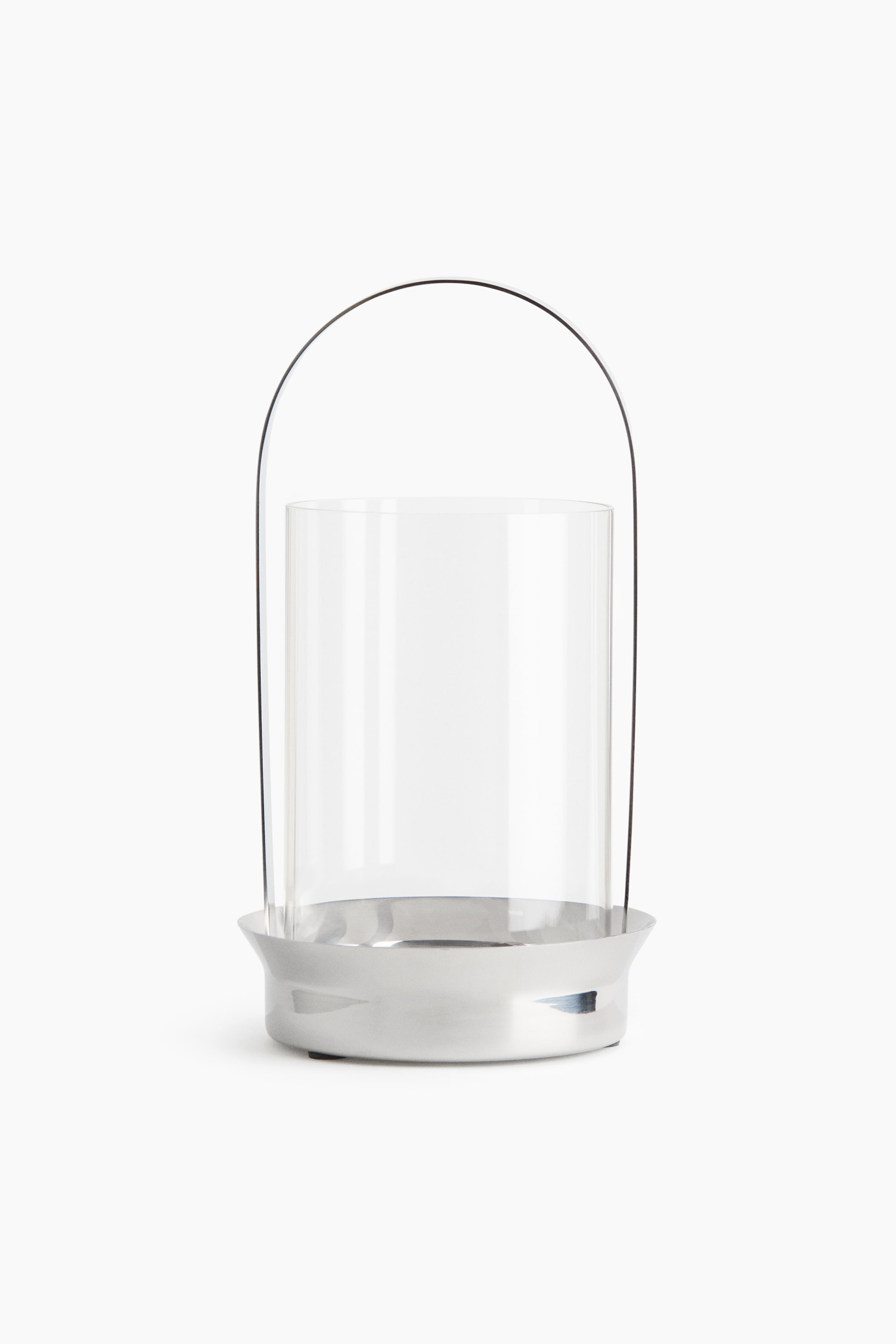 View larger image: The lantern showcases a clear glass cylinder nestled within a sleek, polished silver metallic base. A slender, dark arched handle extends gracefully from the base, curving elegantly over the glass cylinder.