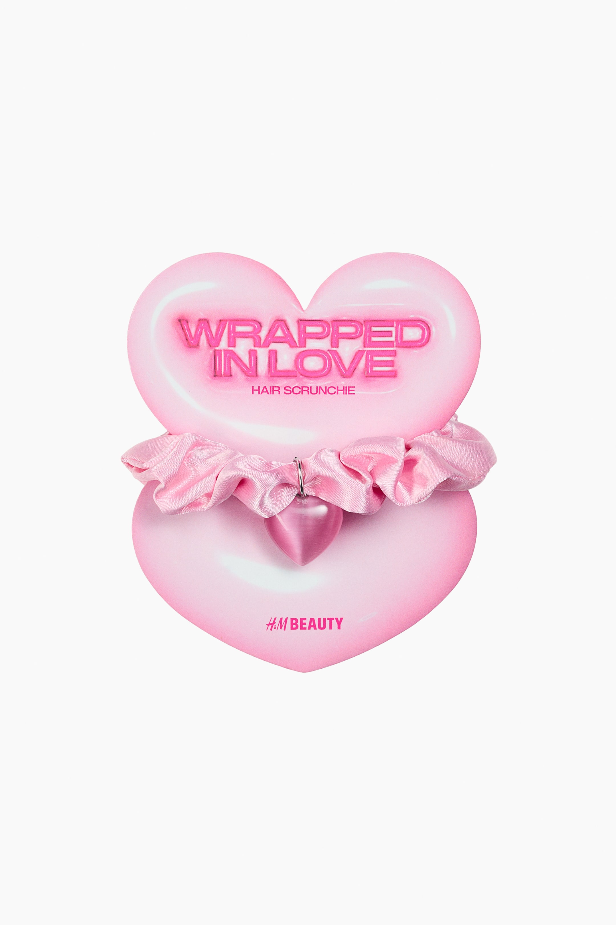 Ampliar la imagen: Un coletero de satén rosa se muestra de frente en una tarjeta de empaque con forma de corazón, envolviéndose alrededor del centro de dos corazones rosas superpuestos. El coletero cuenta con un pequeño dije de corazón rosa sujeto por un anillo metálico, mientras que el empaque muestra el texto "WRAPPED IN LOVE HAIR SCRUNCHIE" en el corazón de arriba y "H.M BEAUTY" en el corazón de abajo.
