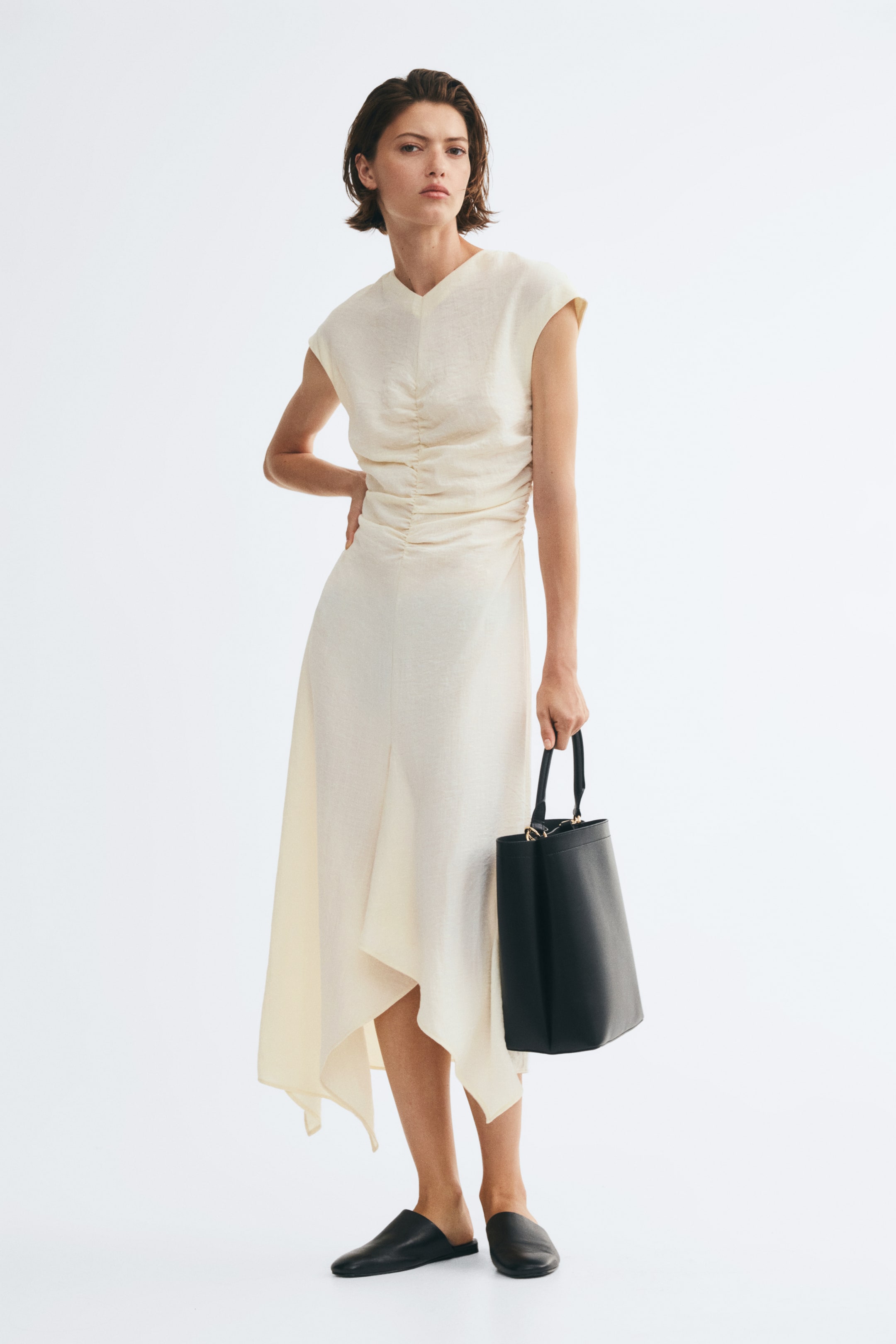 View larger image: The model is wearing a cream-colored, calf-length dress featuring a fitted bodice with central vertical ruching, cap sleeves, and a flared skirt with an asymmetrical handkerchief hem. She is posing with one hand on her hip while carrying a structured black leather tote bag and wearing flat black mules.
