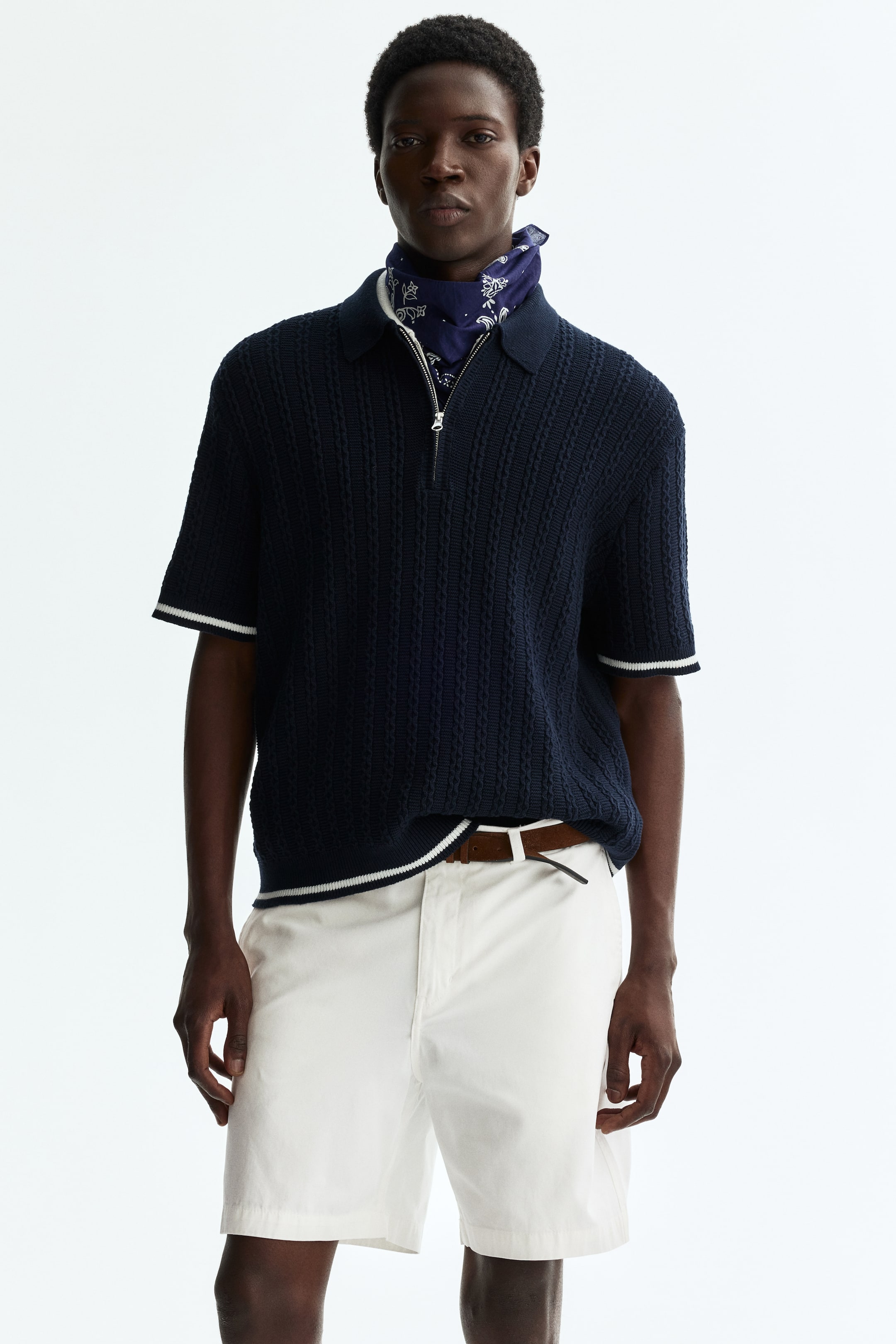 View larger image: A navy blue short-sleeved knitted polo shirt is facing forward, featuring a polo collar, a silver-toned quarter-zip closure, and an open-knit texture throughout. White stripes are adorning the short sleeves' cuffs and the shirt's hem, while a blue patterned bandana is visible under the collar and a white undershirt is showing beneath the partially unzipped front.