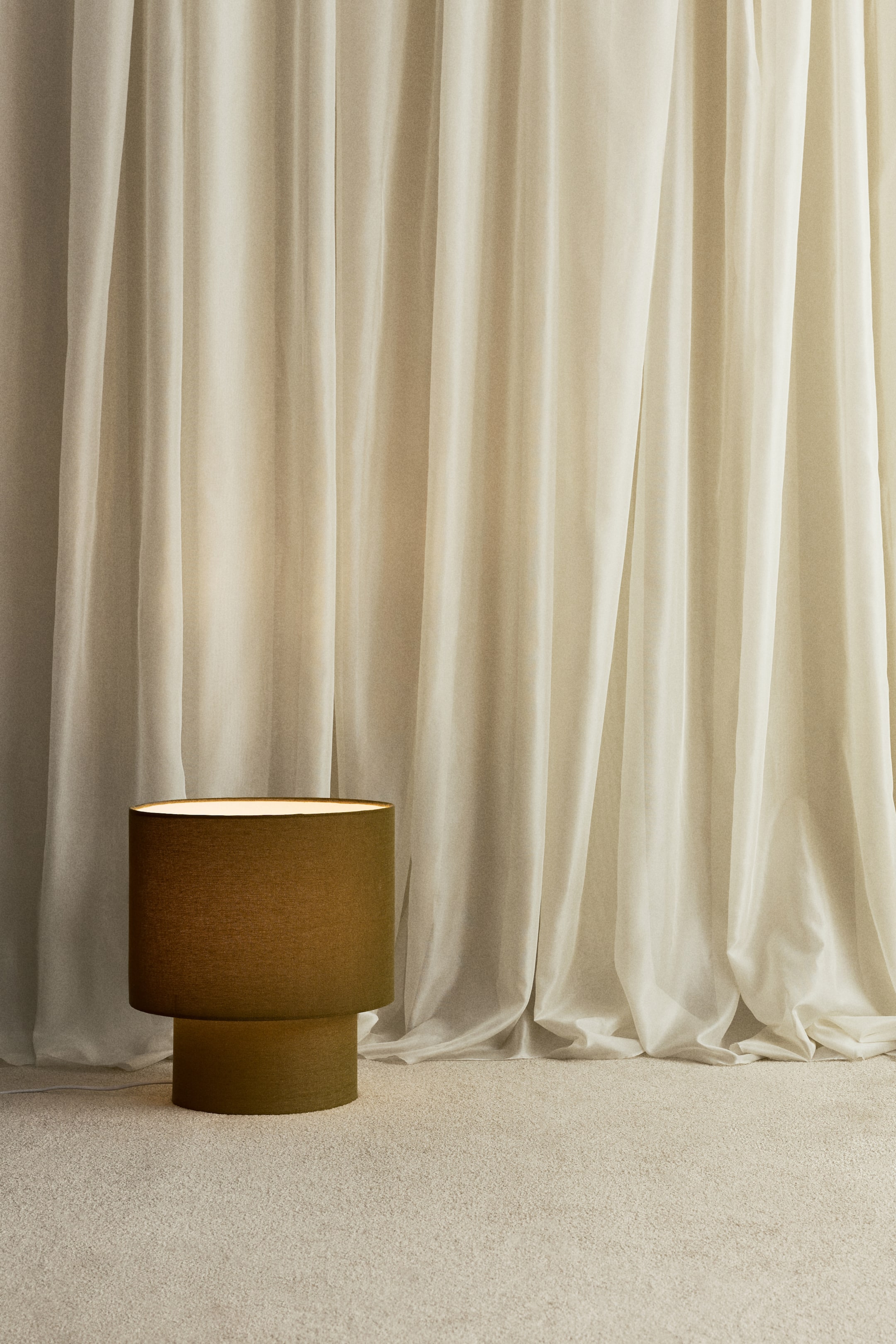 View larger image: A large, front-facing table lamp features a textured, olive-green cylindrical lampshade resting on a narrower cylindrical base, emitting a warm glow from its top.