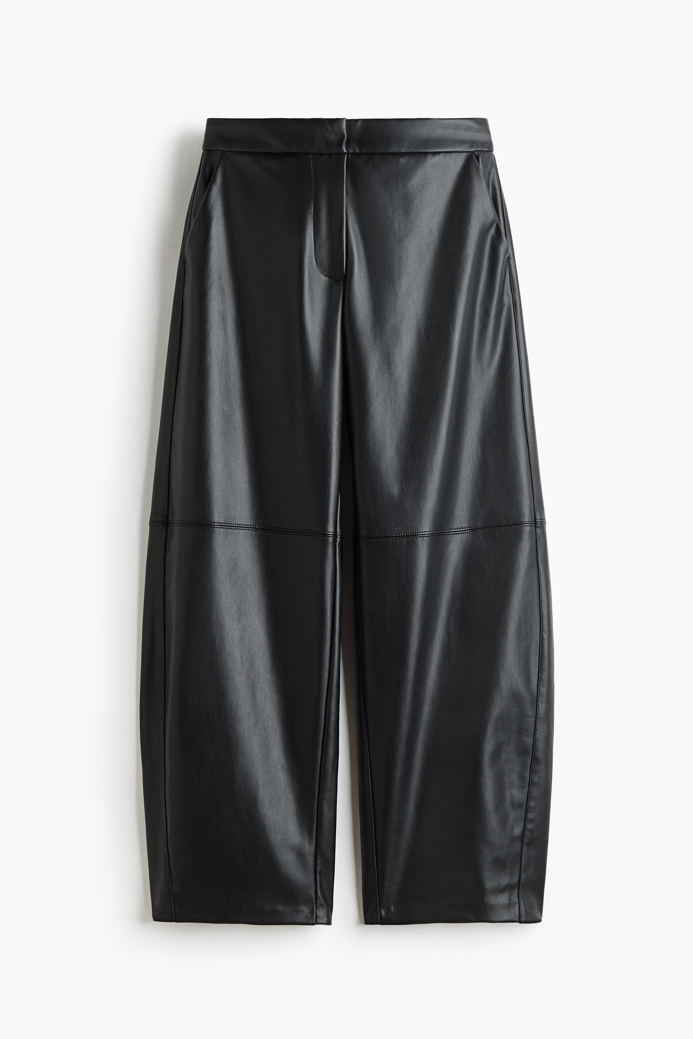 Coated black pants with a shiny finish and balloon legs are being worn by the model, who is sitting on the floor while adjusting the laces of a sneaker. The loose-fitting pants are styled with a long-sleeved, two-tone raglan t-shirt.