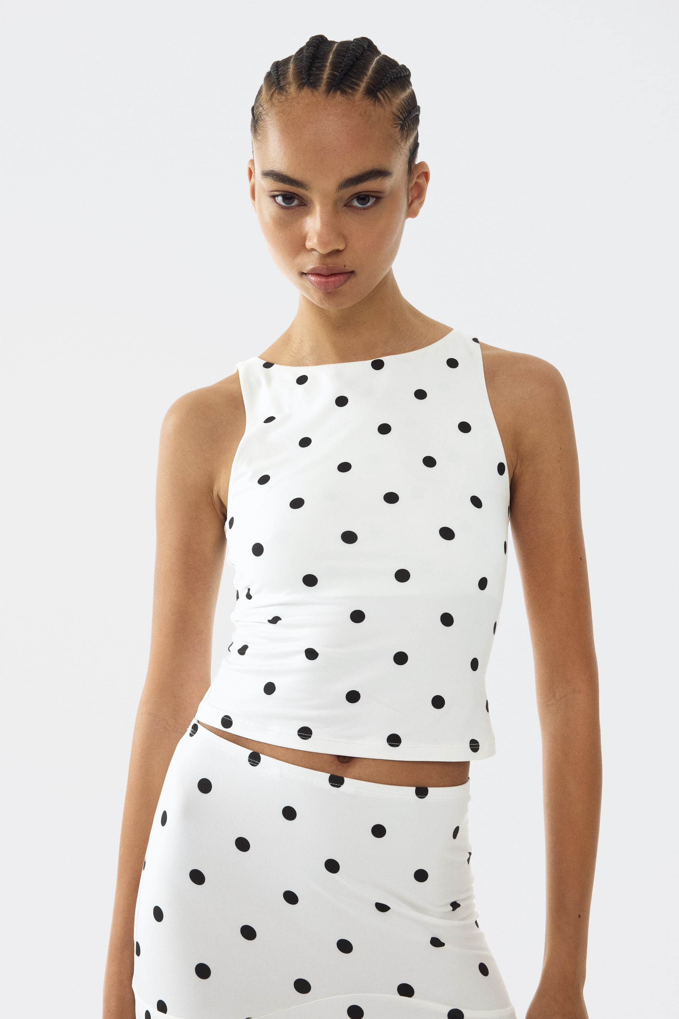 이미지 확대 보기: 앞에서 본 하얀색 민소매 상의는 검은색 물방울무늬가 있으며, 높은 직선형 네크라인과 몸에 딱 맞는 약간 짧은 실루엣을 보여줍니다. 이 상의는 검은색 물방울무늬가 있는 하얀색 치마와 매치되어 있습니다.