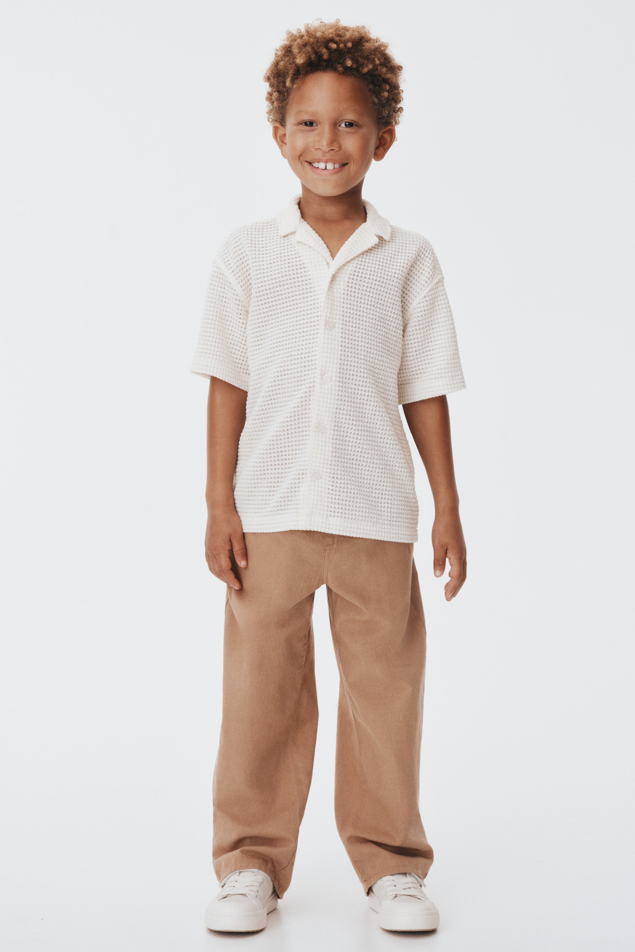 הצגת תמונה גדולה יותר: מראה מקדימה מציג חולצה בצבע לבן-שמנת עם שרוולים קצרים ומרקם סרוג ופל, הכוללת צווארון קמפ פתוח וחזית כפתורים. החולצה משודכת עם מכנסיים בצבע שזוף בהתאמה רגועה, המתקבצים קלות בקרסוליים, ונעלי סניקרס לבנות עם שרוכים נמוכים.