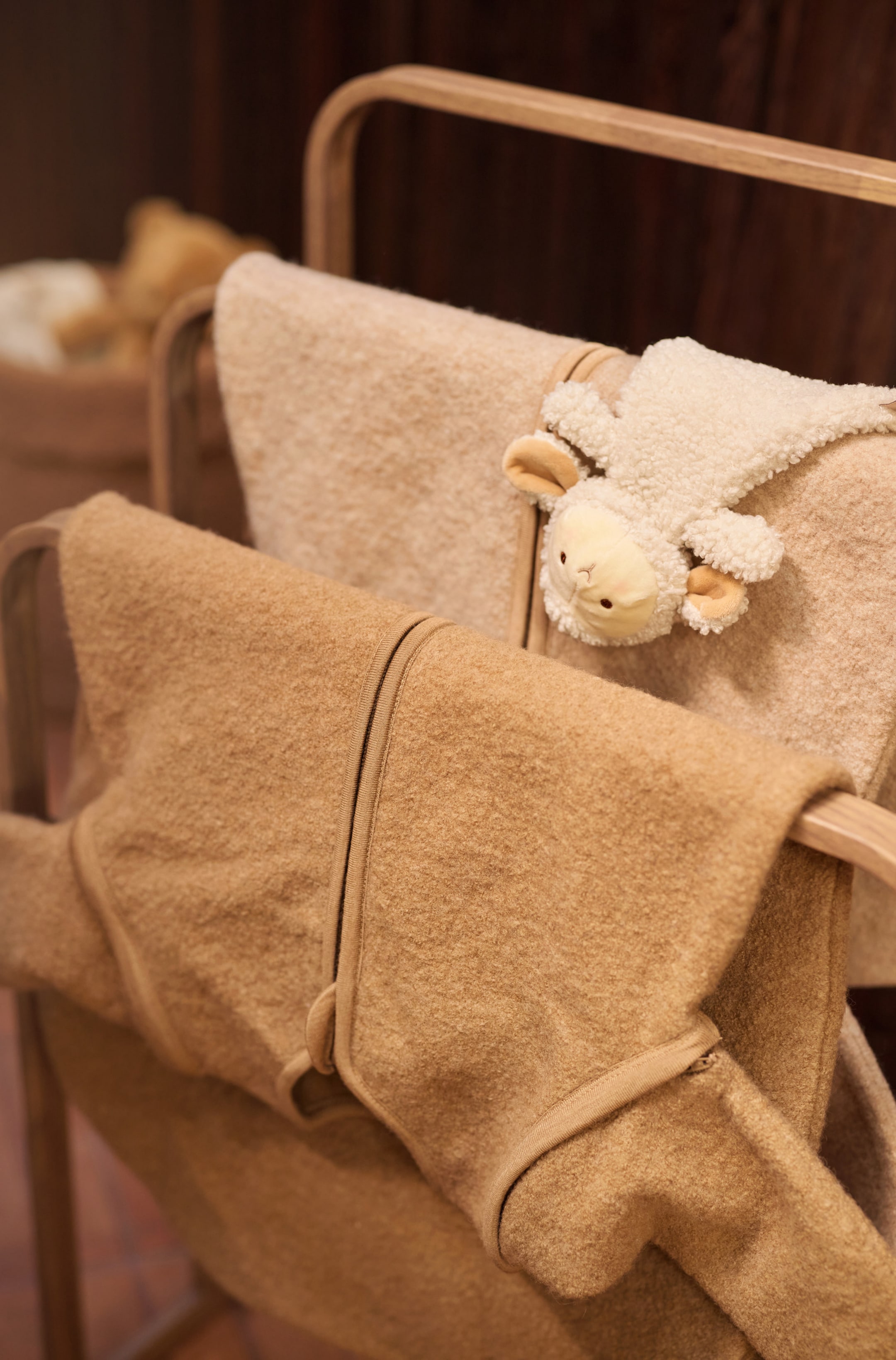 Größeres Bild anzeigen: Der runde Schlafsack mit abnehmbaren Ärmeln – Merino Wool hängt gefaltet über einer Stange und präsentiert seinen weichen, strukturierten Wollstoff in Kamel- und Cremetönen. Die kamelfarbenen Ärmel im Vordergrund zeigen Kanten mit Reißverschluss und passend eingefasste Abschlüsse, während die cremefarbene Variante dahinter mit einem kleinen, kuscheligen Schaf-Spielzeug darüber drapiert ruht.