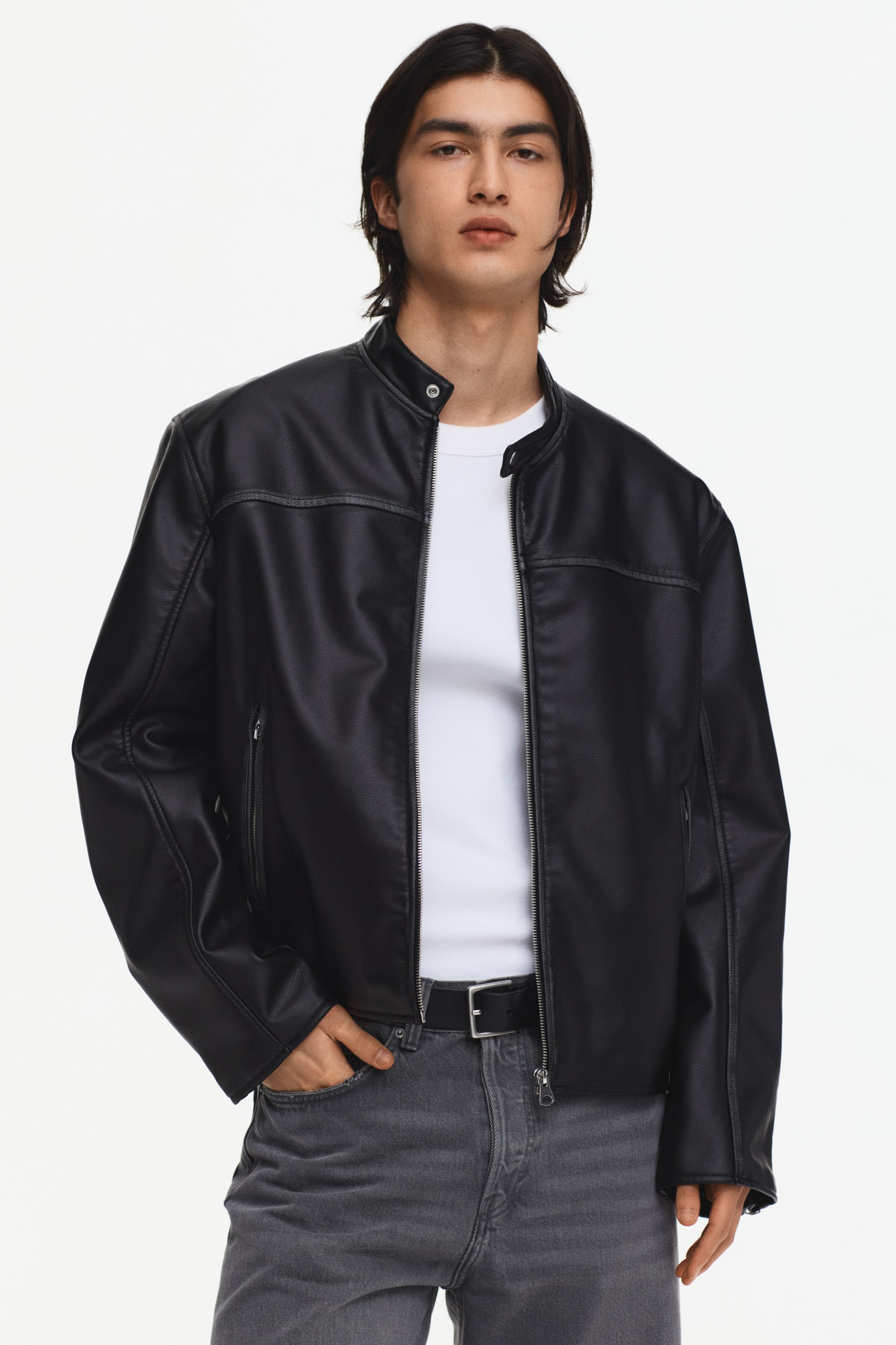 Größeres Bild anzeigen: Eine schwarze Kunstlederjacke wird von vorne präsentiert. Sie verfügt über einen Stehkragen mit Druckknopfverschluss, einen teilweise geöffneten mittigen Frontreißverschluss, der ein weißes T-Shirt darunter zeigt, und eine vertikale Reißverschlusstasche auf der linken unteren Vorderseite.