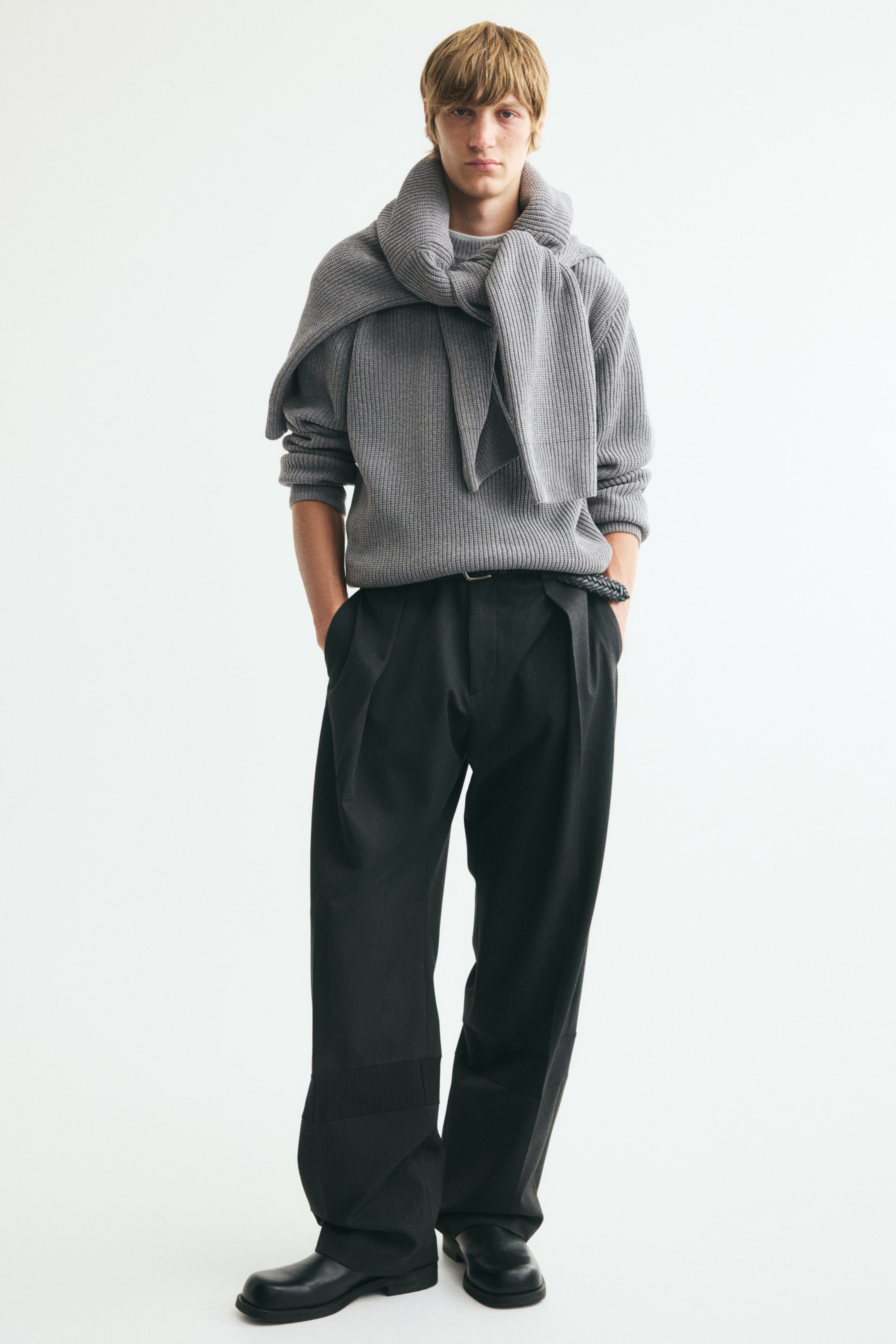 View larger image: A grey rib-knit jumper with a round neckline, dropped shoulders, and long sleeves is worn by the model. This relaxed-fit jumper is styled with a matching grey rib-knit scarf draped over the shoulders and tied at the front.
