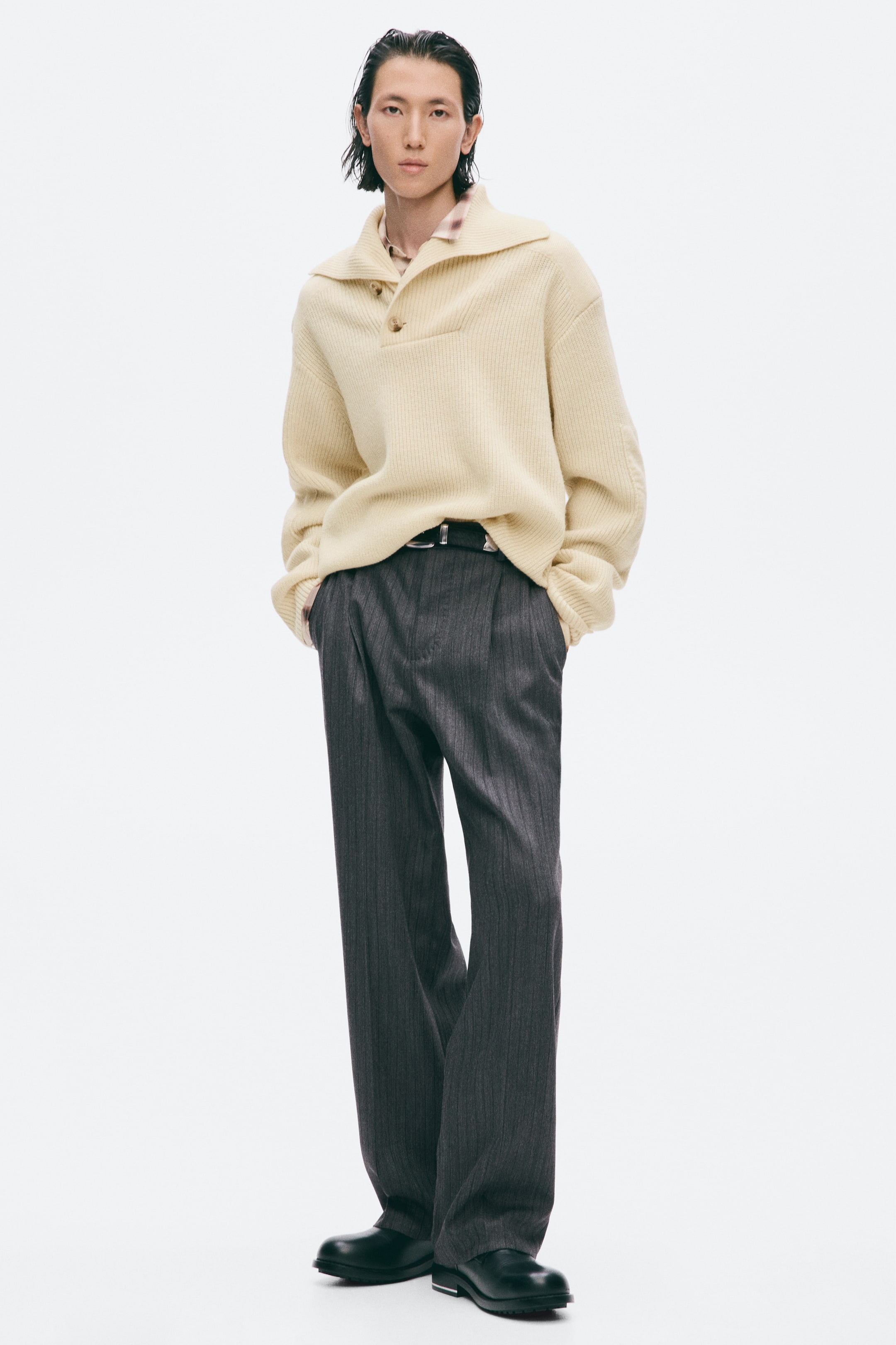 Ver imagem maior: Uma camisola de malha canelada, de cor creme, com gola larga abotoada, está a ser usada pela modelo por cima de uma camisa com padrão e gola. A camisola está ligeiramente metida pela parte da frente dentro de calças cinzento-escuras às riscas diplomáticas.