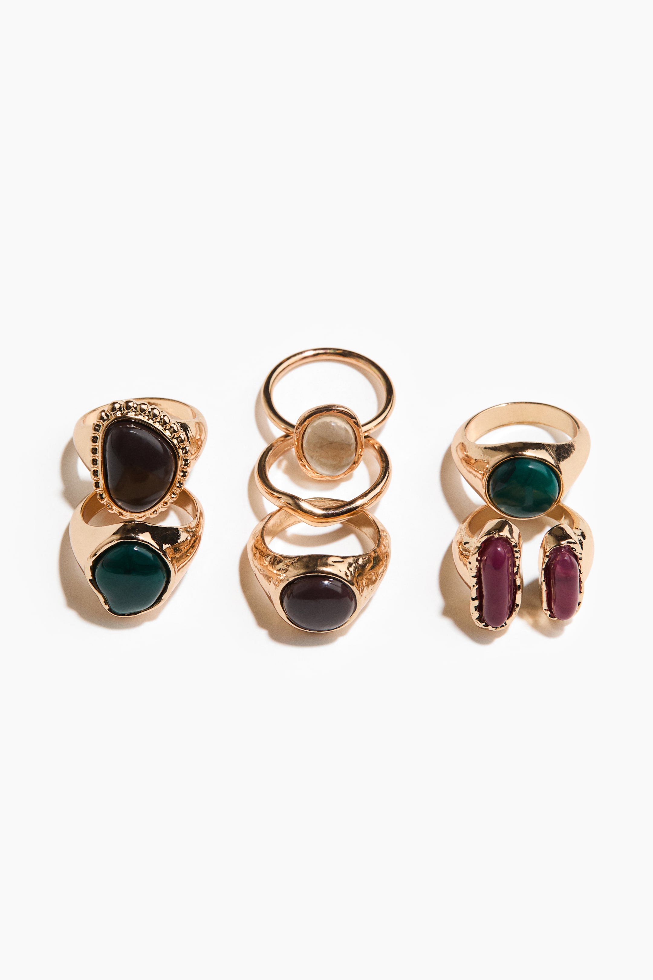 View larger image: Three sets of gold-toned rings are on display, each featuring various coloured stones. The designs include rings with irregular dark brown, oval dark green, oval translucent light grey, and rectangular dark red stones, with some bands showcasing textured or beaded detailing.