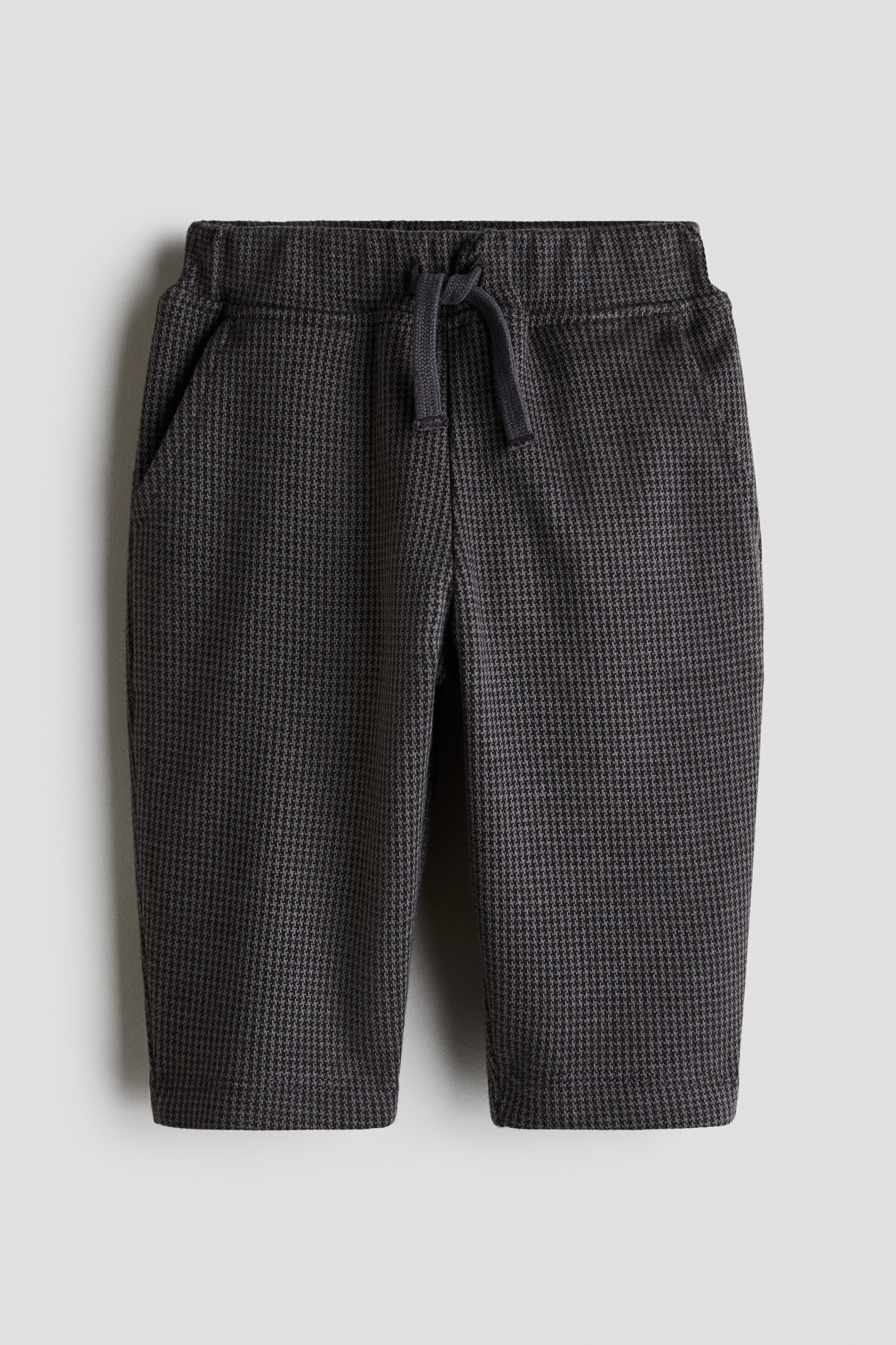 These dark grey trousers in textured jacquard jersey are featured from the front on a toddler, showing a relaxed, straight-leg fit. The trousers are styled with a green and red plaid shirt, a beige cable-knit cardigan, and dark brown loafers.