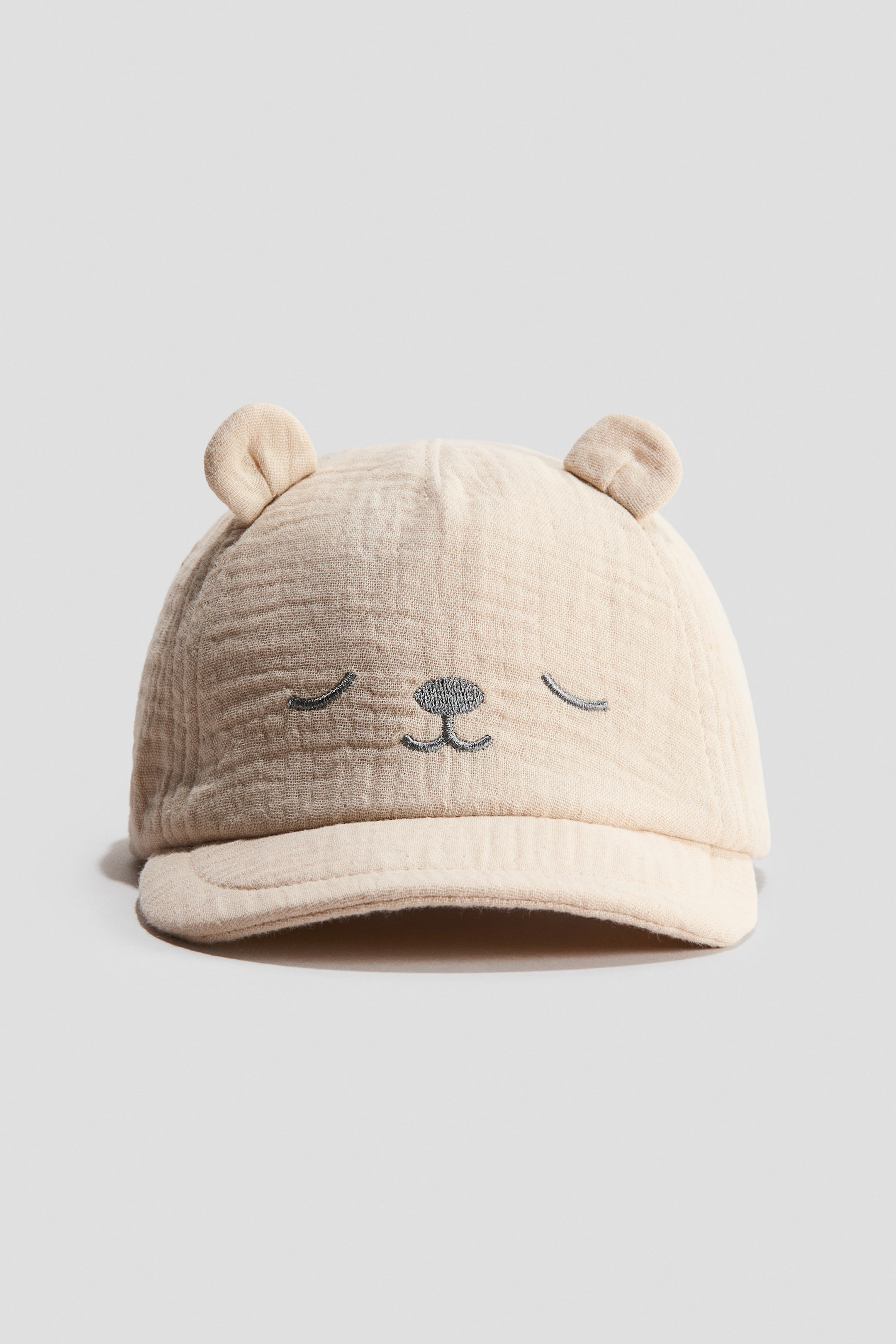 View larger image: "A soft-peak muslin cap is facing front, showcasing its light beige crinkled texture, two small rounded ears on top, and a dark grey embroidered sleeping face design."