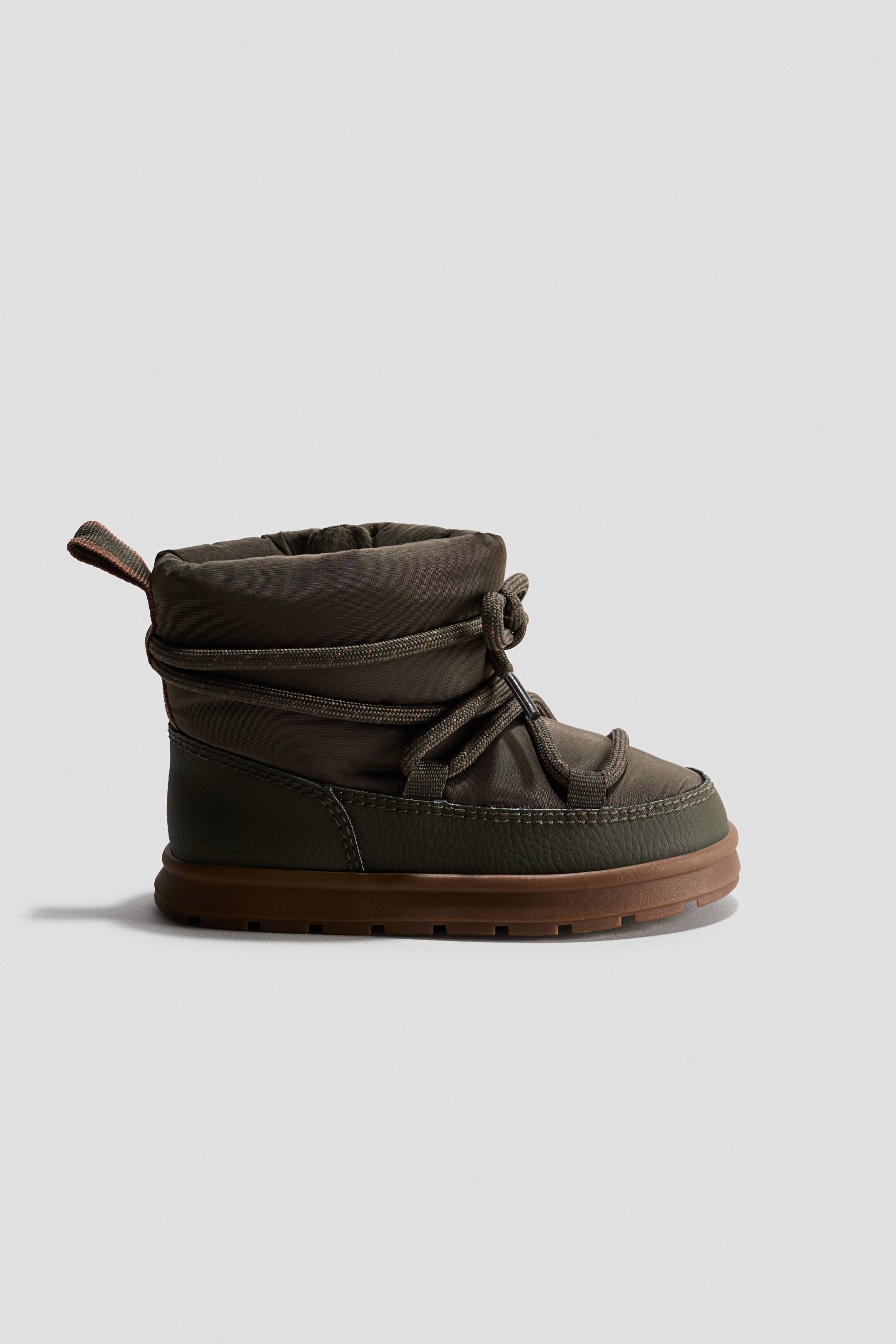 View larger image: An olive green winter boot is shown in side profile, featuring a padded nylon upper with a matching textured mudguard. Thick rope laces wrap around the ankle shaft, and a chunky brown rubber sole completes the design.