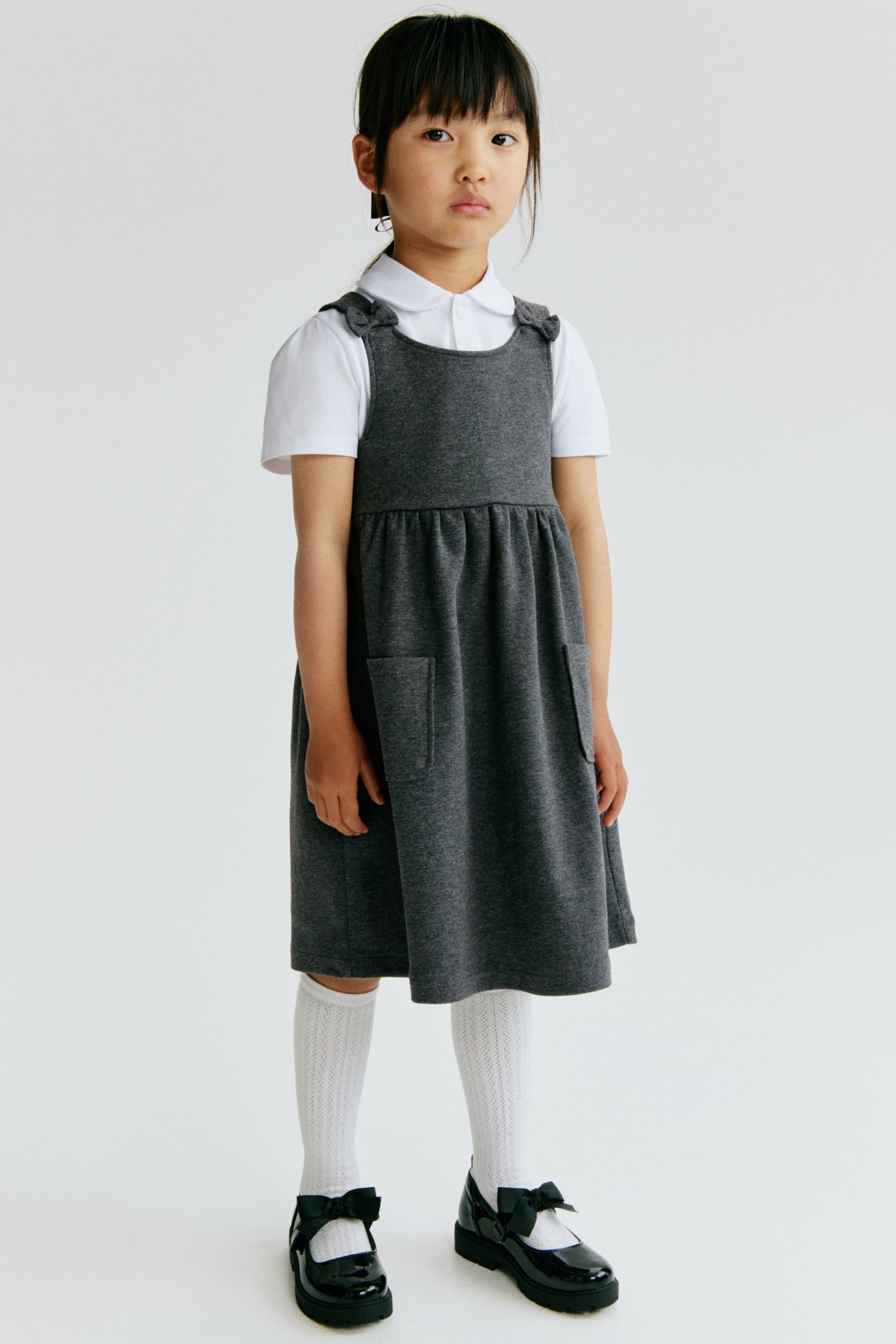 View larger image: A dark gray pinafore dress features shoulder straps tied into bows and two front patch pockets, with the skirt gathered at the waist and falling above the knees. It is layered over a white short-sleeved polo shirt.