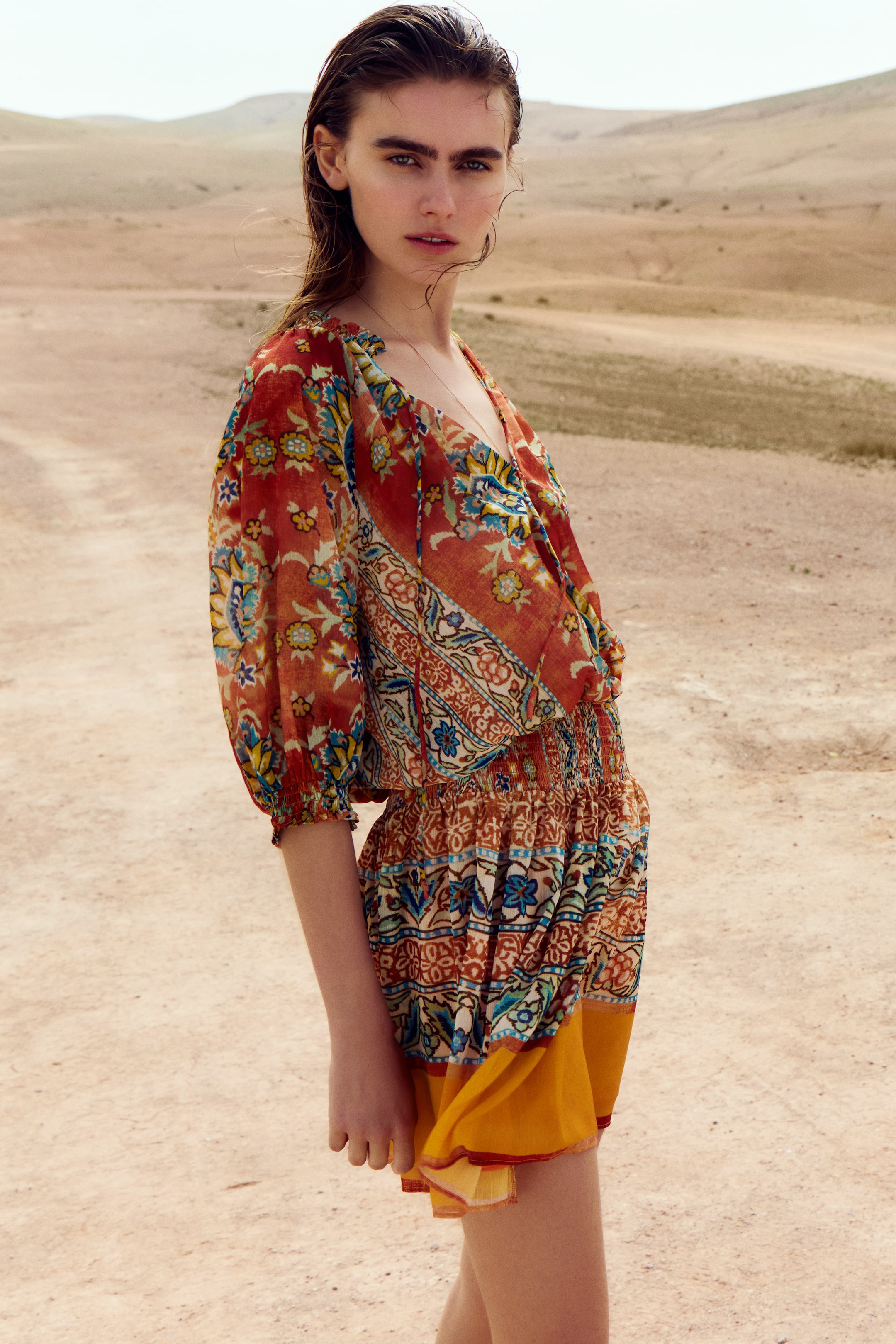 Größeres Bild anzeigen: Ein kurzes, oversized Tunika-Kleid aus gewebtem Stoff mit einem aufwendigen Muster wird vom Model getragen. Es verfügt über einen runden, gesmokten Ausschnitt mit einem V-förmigen Schlitz und Spaghettibändern, kurze, voluminöse Raglanärmel mit Smokdetails an den Bündchen, einen gesmokten Taillenbund und einen ausgestellten Rock, der teilweise mit gelbem Stoff gefüttert ist.