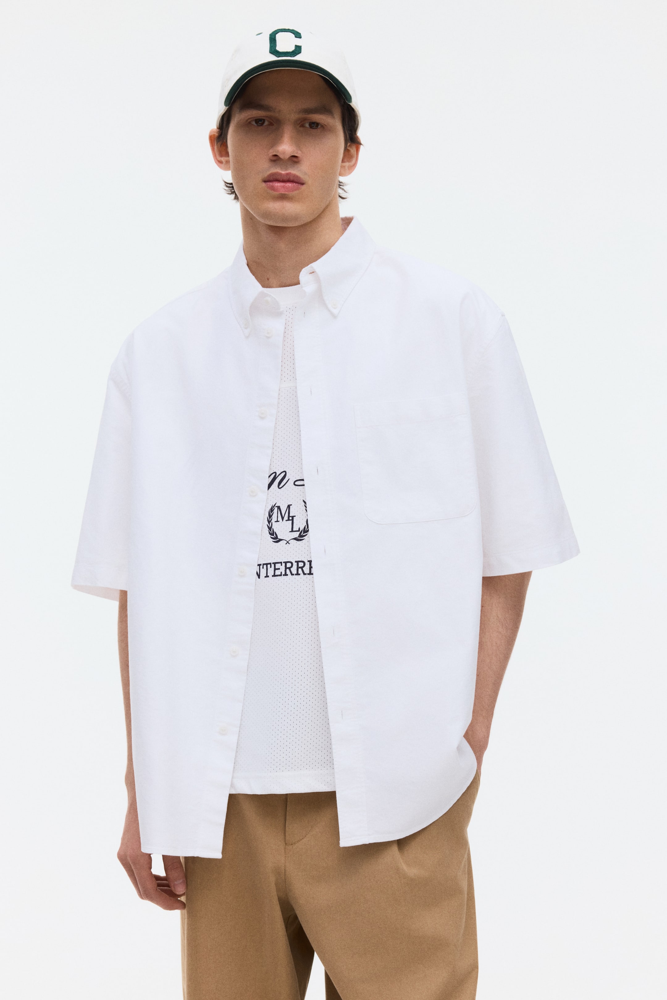 Ver imagen más grande: La camisa blanca de manga corta presenta un cuello abotonado, cierre frontal con botones y un único bolsillo parche en el pecho izquierdo. Ofrece un ajuste relajado y se lleva abierta sobre una camiseta de malla blanca.