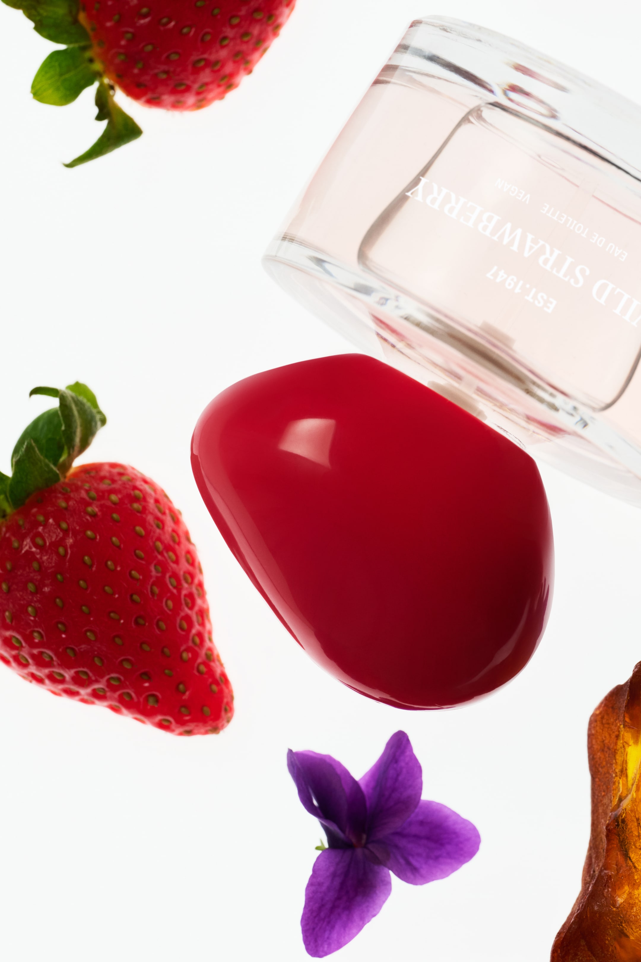 Visa större bild: En klar rektangulär glasparfymflaska ligger diagonalt, vinklad från övre högra hörnet till nedre vänstra hörnet. Den visar ljusrosa vätska inuti och har texten "WILD STRAWBERRY EST.1947 EAU DE TOILETTE VEGAN" i vit text på sidan, medan den vilar på en glansig, slät, ljusröd, avlång formad kork.