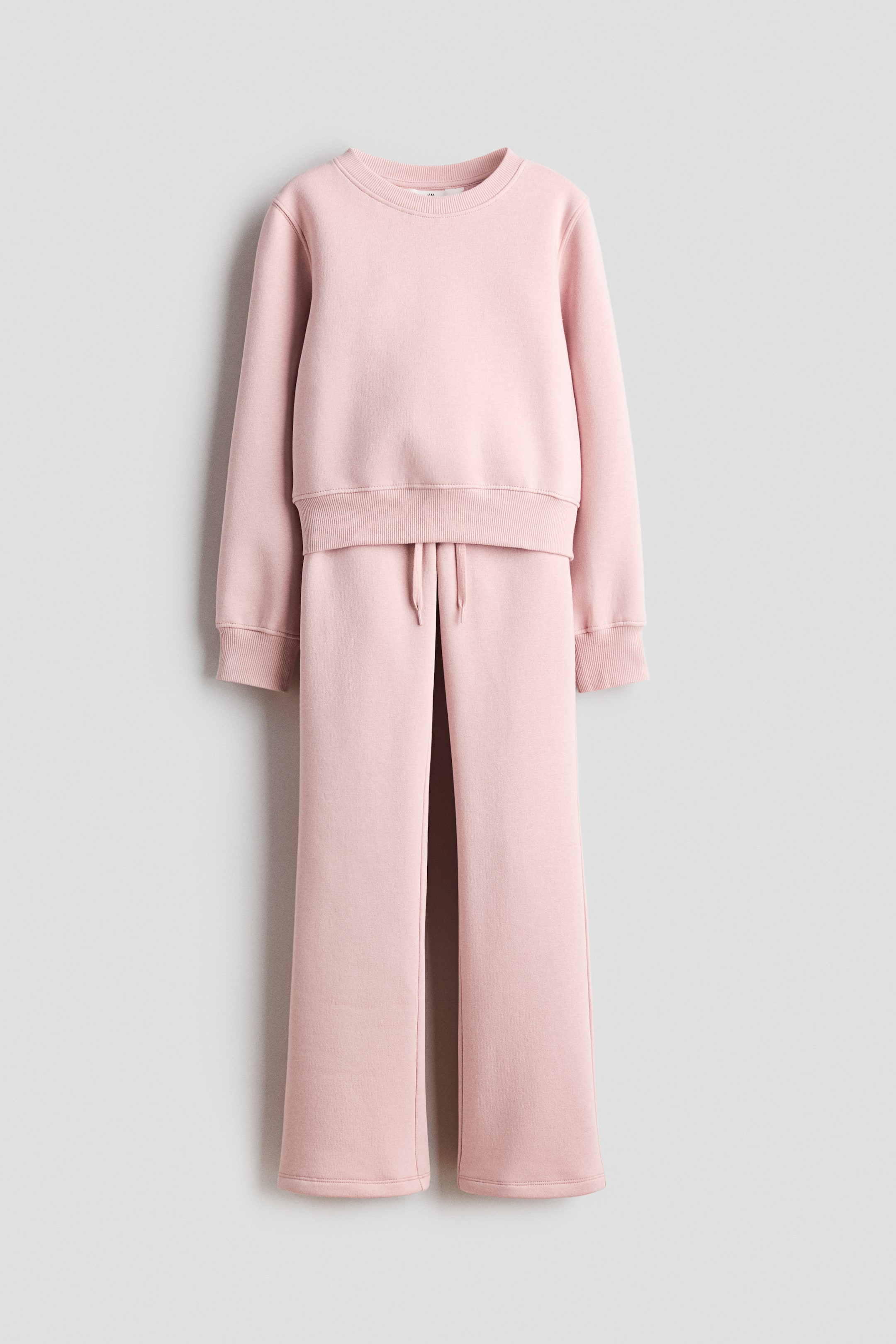 Ver imagen más grande: Un conjunto de sudadera rosa, confeccionado en tejido de sudadera, está colgado de frente. La parte superior de manga larga presenta un cuello redondo con ribete acanalado y puños y dobladillo acanalados, mientras que los joggers a juego muestran una cintura elástica con cordón visible y piernas anchas y rectas.