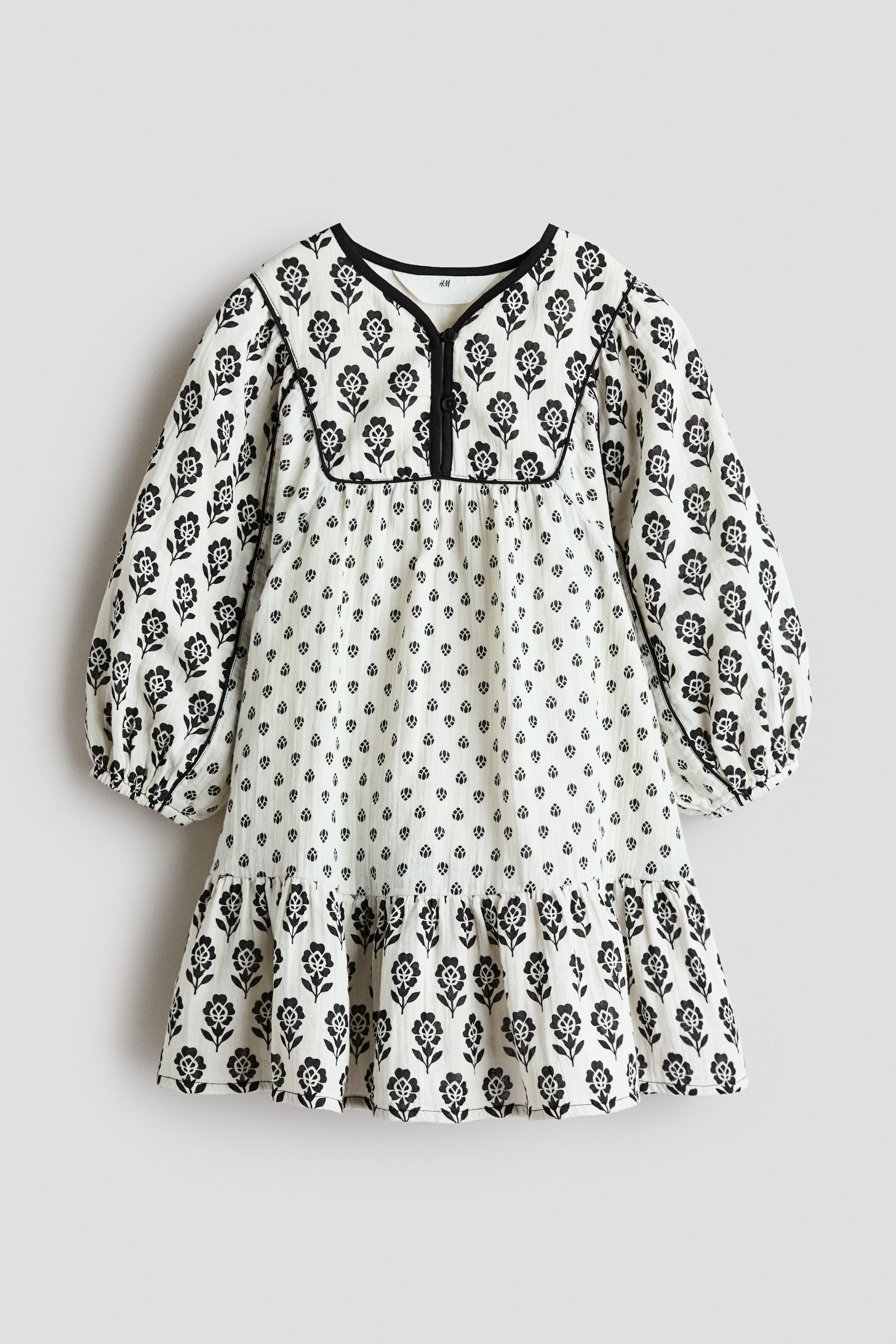 View larger image: A white A-line dress is showcasing its front, adorned with black floral patterns of varying densities. It is crafted with a V-neck and features two button-and-loop fasteners on a square yoke, from which gathers elegantly fall. Long balloon sleeves with elasticated cuffs extend gracefully, and a gathered tier forms the hem.