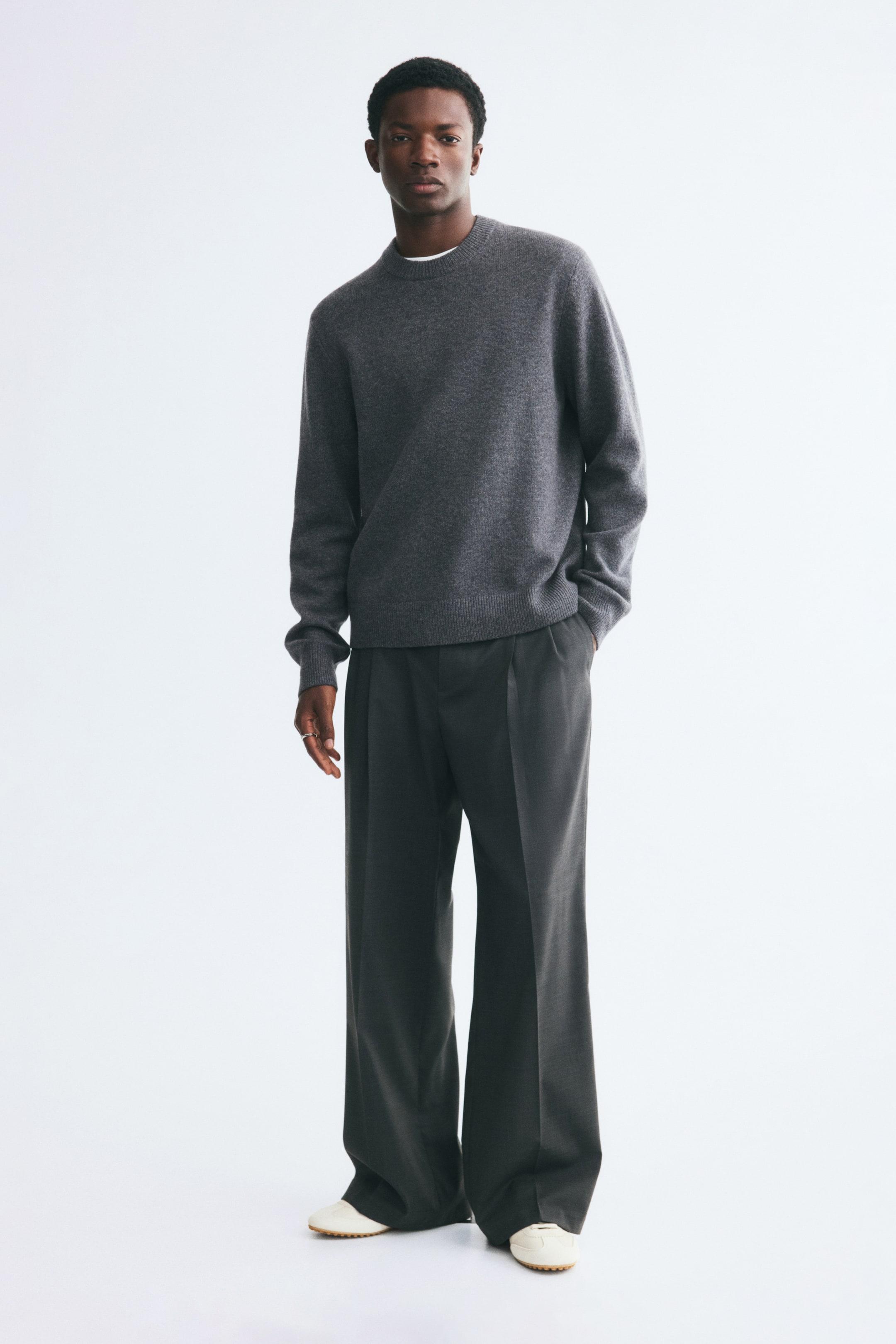 View larger image: The model is wearing a gray knitted wool sweater with a round ribbed neckline, long sleeves, and ribbed cuffs and hem. The sweater is layered over a white collar and paired with wide-leg dark gray trousers.