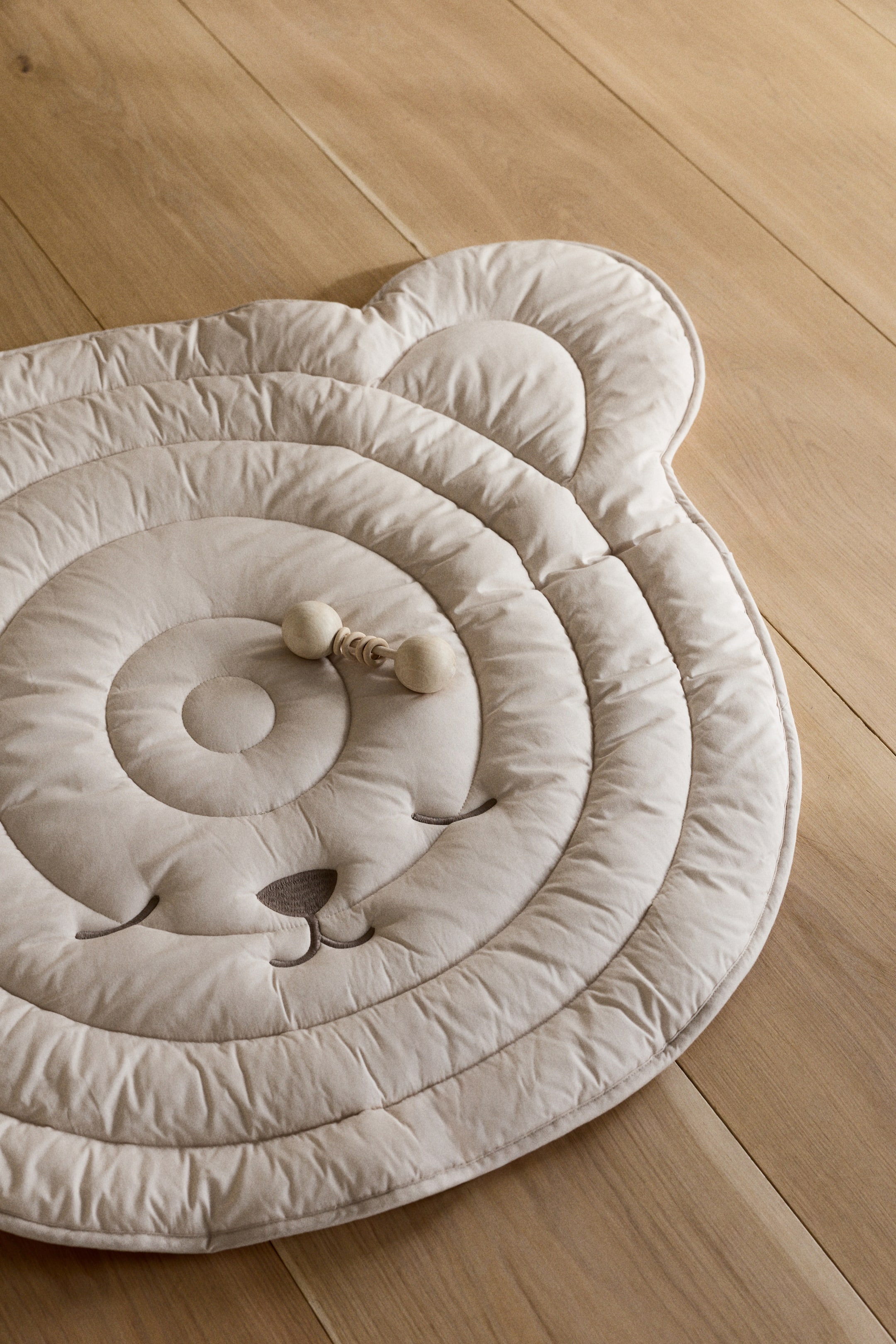 Agrandir l'image: Un tapis de jeu en forme d'ours de couleur claire présente des coutures matelassées formant des cercles concentriques sur sa surface supérieure, ainsi qu'un visage brodé. Un hochet en bois repose en son centre.