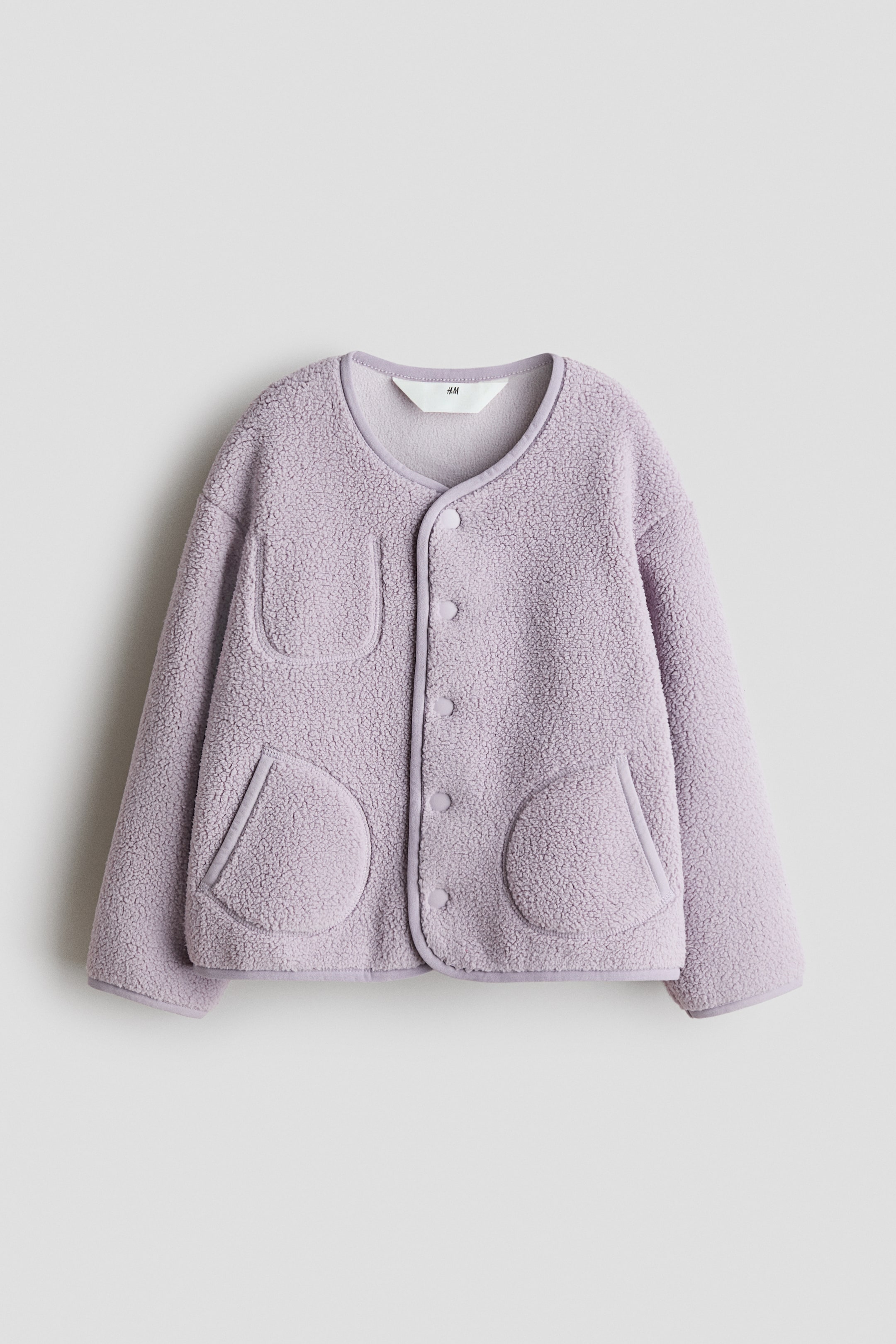 拡大画像を表示: ラベンダーのフリースジャケットは、前面から提示され、テクスチャーのあるフリース素材とラウンドネックラインが特徴です。ボタンは前面に5つの同色ボタンと、滑らかな生地でトリムされたパッチポケットが3つデザインされています。ひとつは左上にある長方形のポケットで、残りの2つは左下と右下にある丸いポケットです。