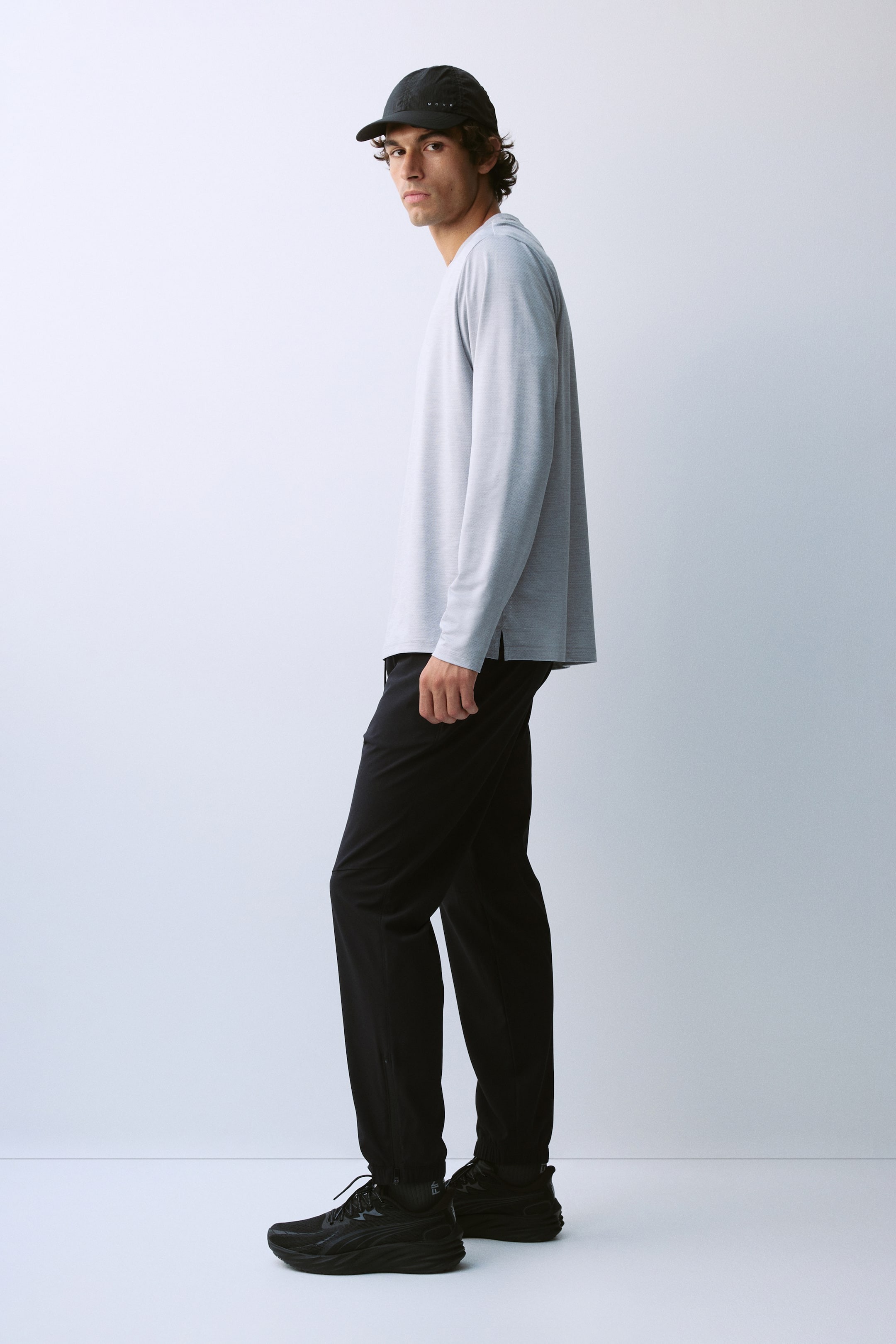 View larger image: A light grey long-sleeve top featuring a crew neck and side slits at the hem is worn by the model. The top is styled with black trousers, black sneakers, and a black baseball cap.