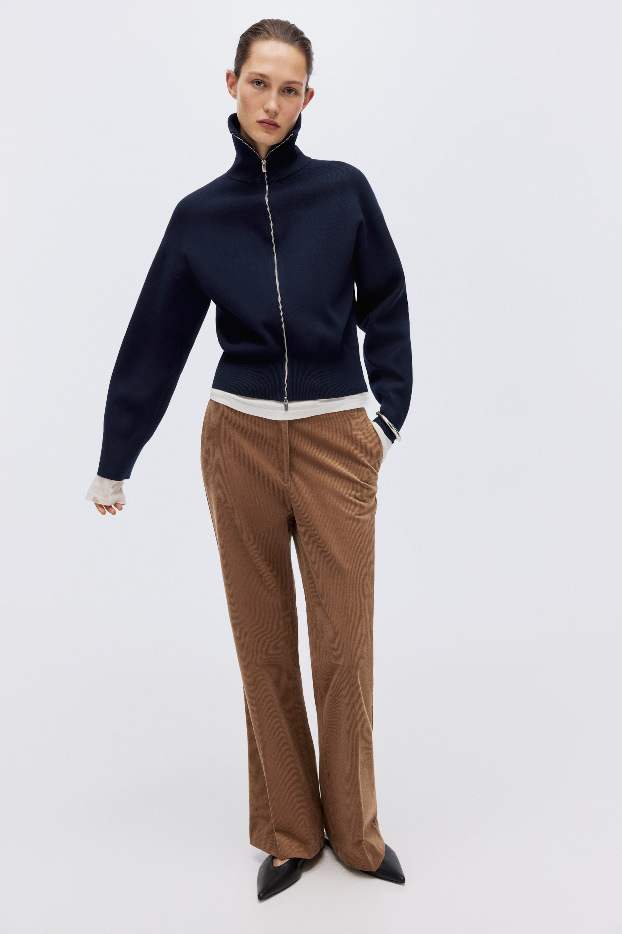 拡大画像を表示: モデルは、ファインニットのネイビーブルーのカーディガンを着用しています。襟は高さのあるリブ仕様で、ドロップショルダー、袖口と裾にはリブが施されています。ツーウェイジップは下側から一部開けた状態で、裾と袖口から白いトップスを重ね着しているのが見えます。