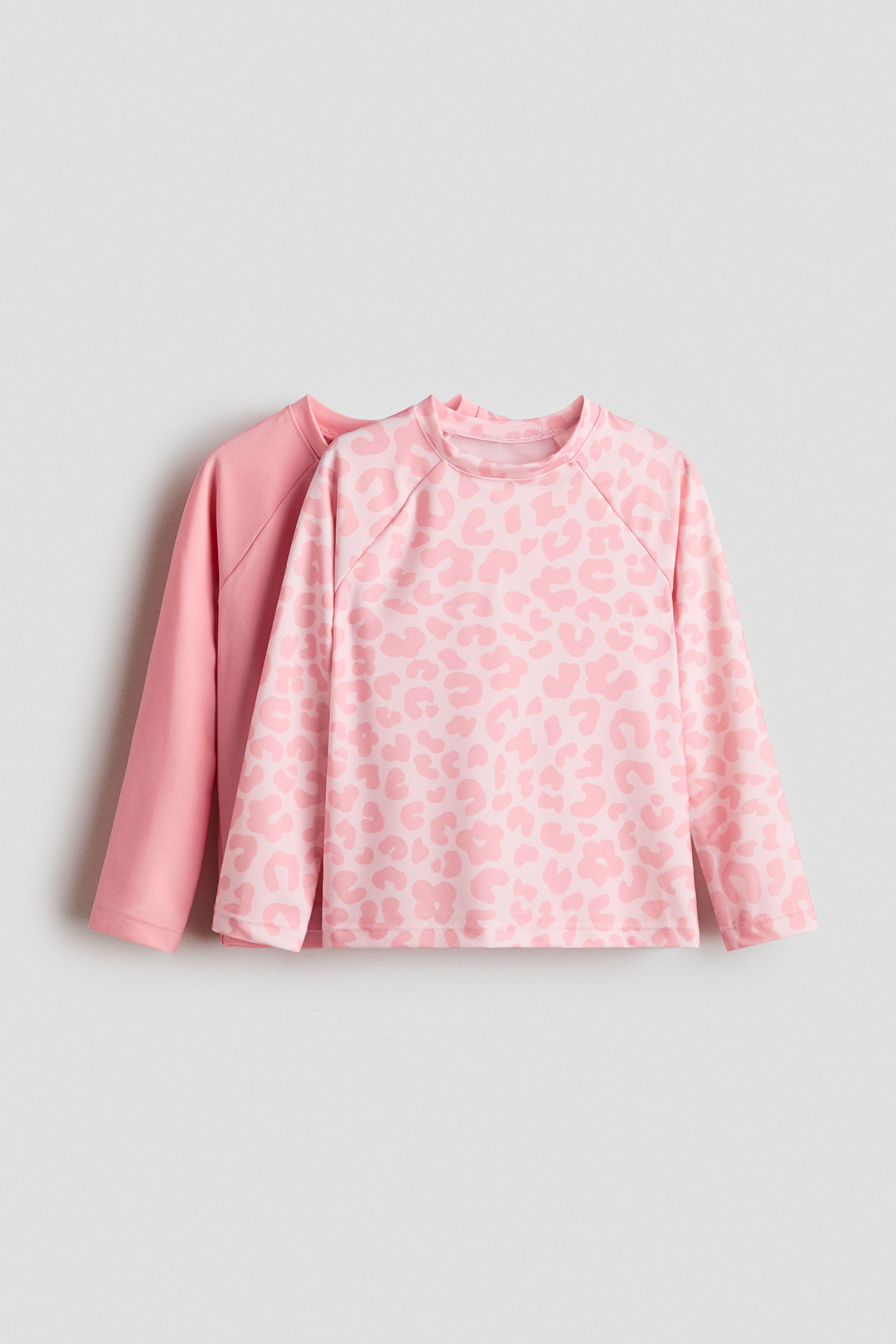View larger image: Two long-sleeve tops are shown, one in front of the other. The front top features a light pink leopard print on a pink base, a crew neck, and raglan sleeves. The top behind displays a solid pink color, also with a crew neck and raglan sleeves.