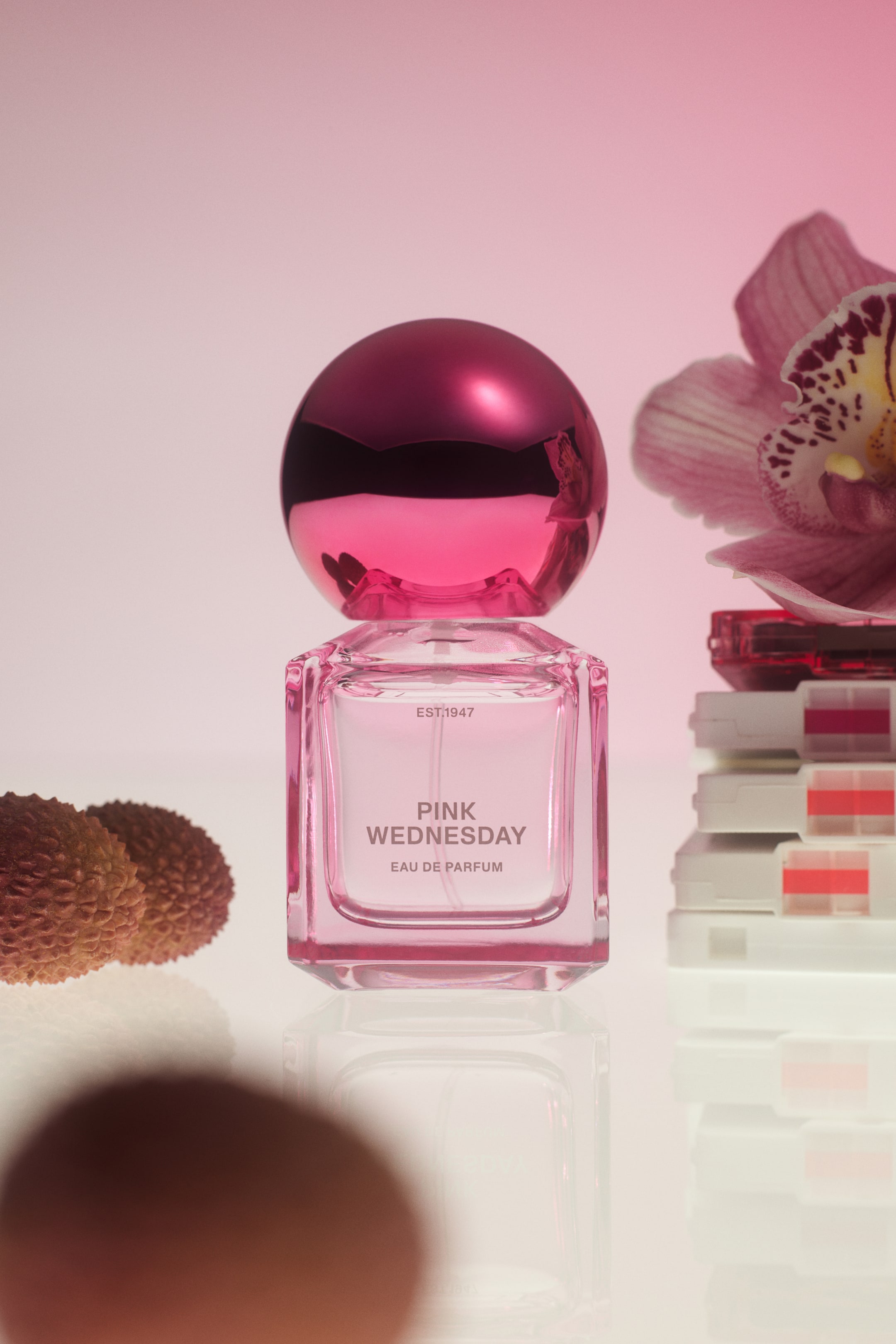 Zobraziť väčší obrázok: Štvorcová sklenená flakónová fľaštička s jemne ružovým odtieňom, označená „PINK WEDNESDAY EAU DE PARFUM“, stojí vzpriamene a je zakončená guľovitým kovovým ružovým uzáverom. Fľaštička je umiestnená na odrazovej ploche spolu s plodmi liči a ružovým kvetom orchidey položeným na kôpke kaziet, čím vizuálne predstavuje kľúčové tóny vône.