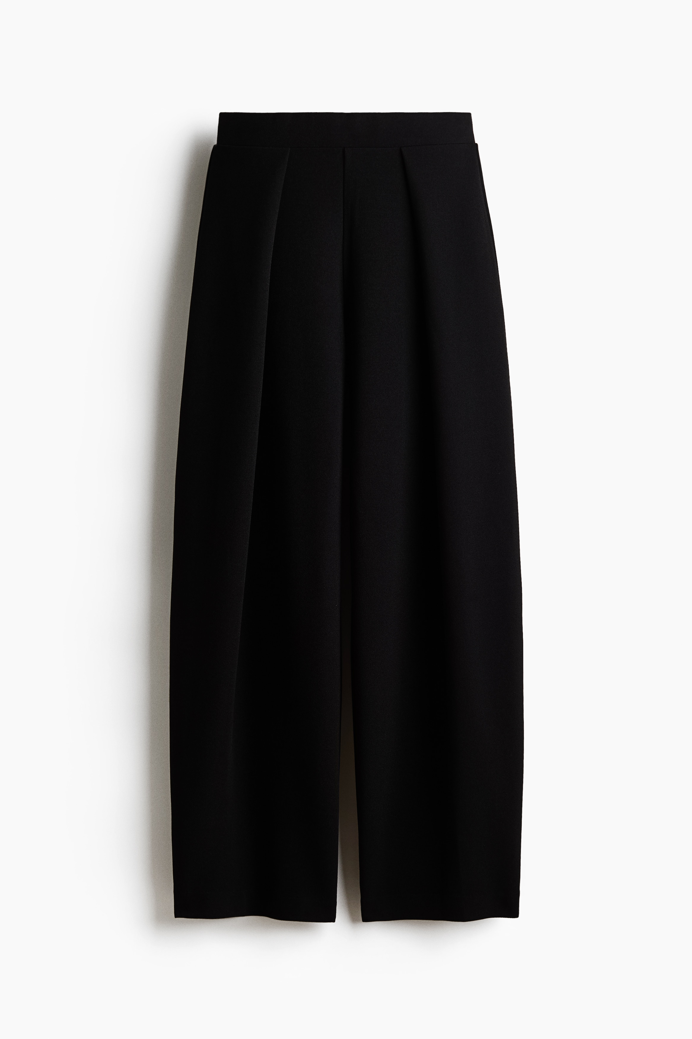 Loose fit pants in black ponte fabric are being worn by the model, featuring a high waist and balloon legs with draped wrap sides. The pants are styled with a cropped top and an open, striped shirt.