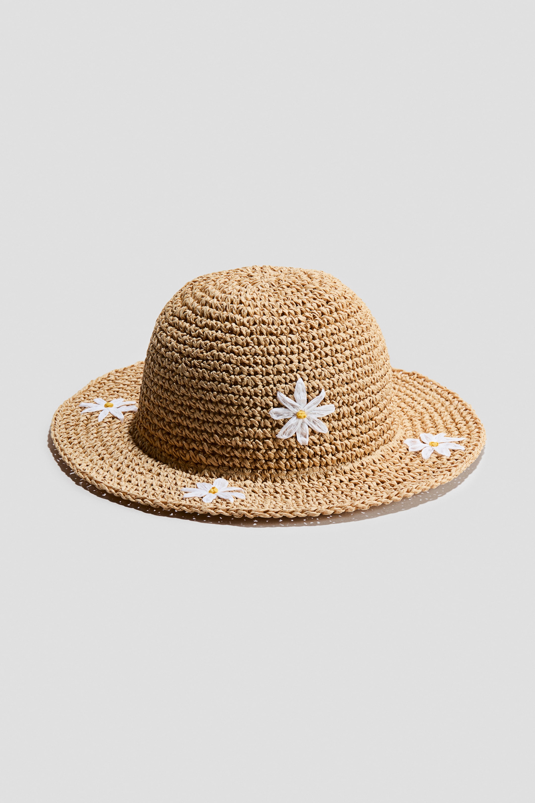Ingrandisci l'immagine: Un cappello di paglia intrecciata presenta una corona arrotondata e una tesa larga con una trama aperta visibile. Applicazioni di margherite bianche con centri gialli decorano la tesa e la parte inferiore della corona.