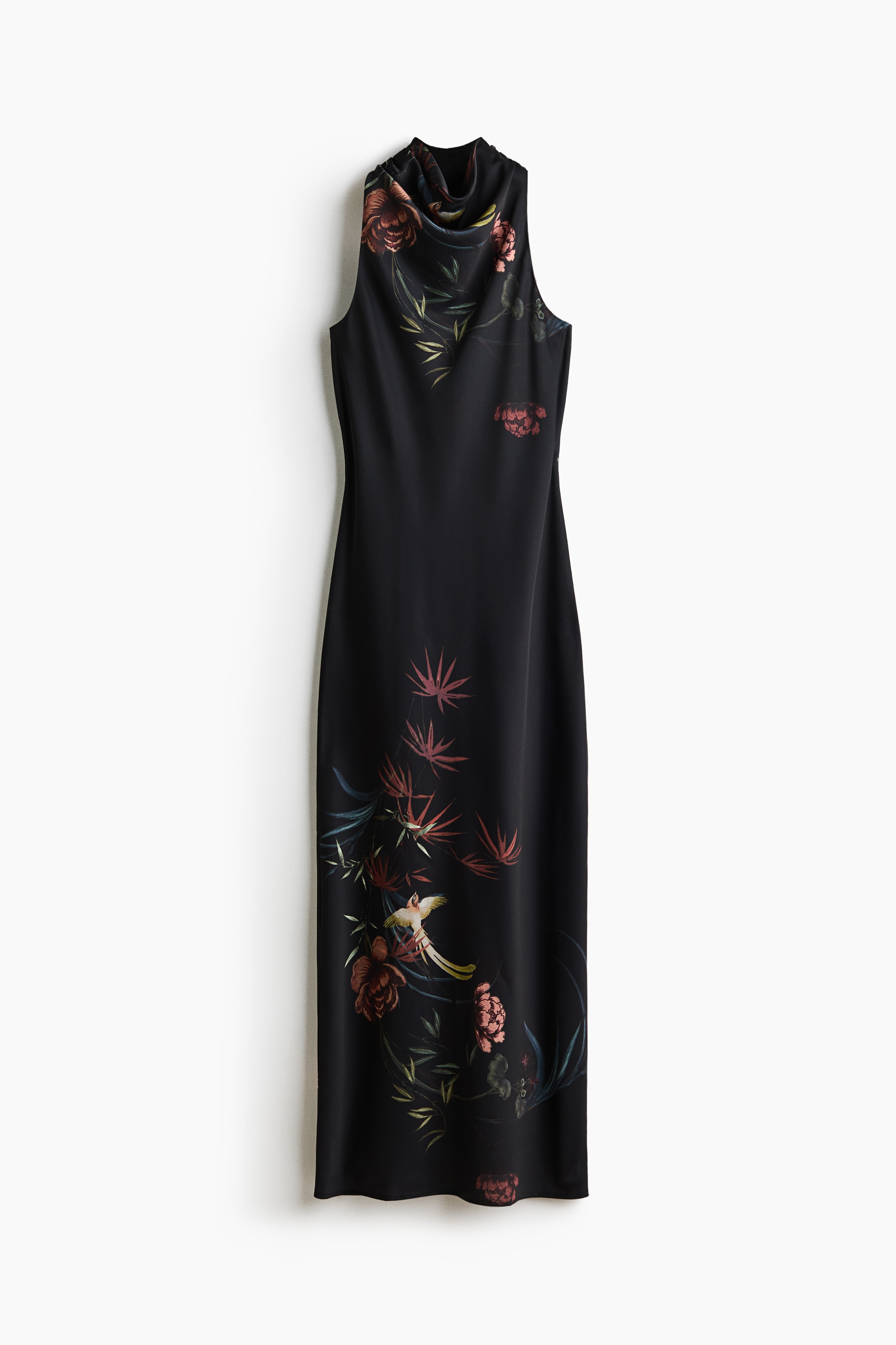 A long, sleeveless black dress featuring a botanical and bird print on the upper back is being worn by the model, who is standing with her back partially turned. The woven dress has a subtle sheen, shaping seams at the waist, and a slit at the back hem.