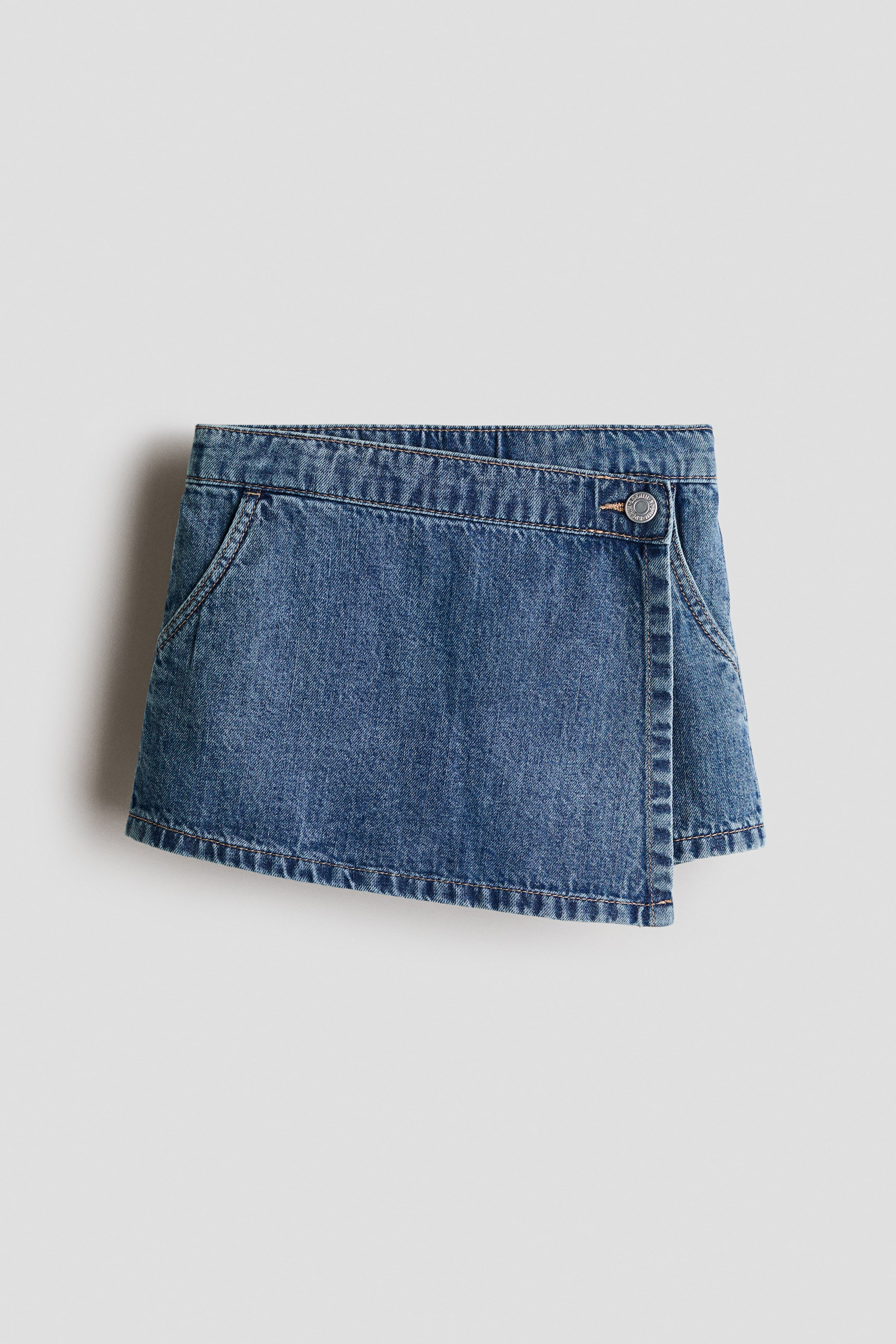 View larger image: A medium wash blue denim skort features an asymmetrical wrap-style front, fastened by a single silver-toned button on the right side, showcasing two angled front pockets with visible contrast stitching.
