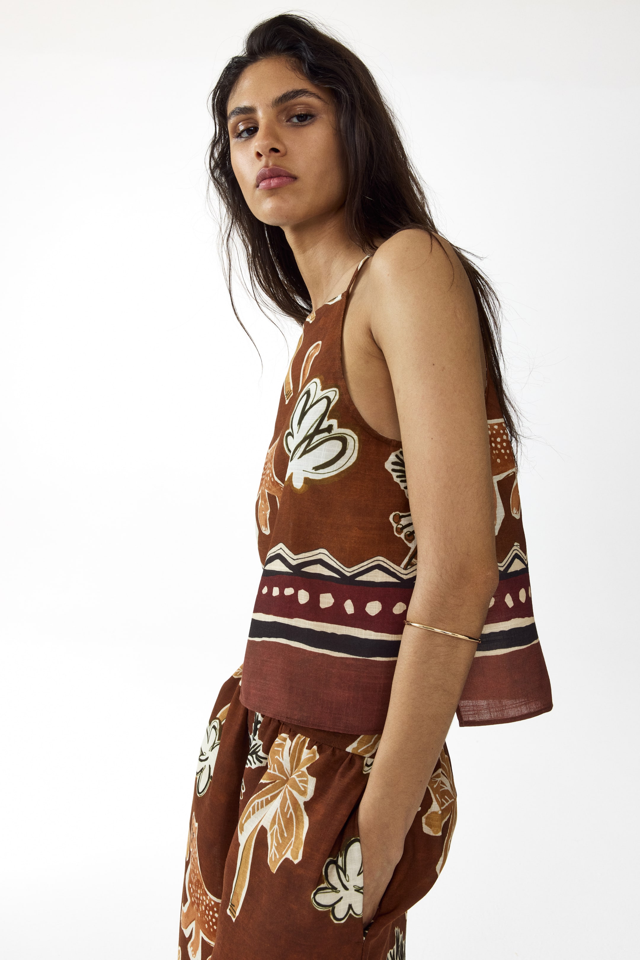 Ver imagem maior: Um top sem mangas em tom de terracota e calças a condizer estão virados para uma vista lateral de três quartos, exibindo um padrão botânico estilizado em creme, branco e bege. O top apresenta alças finas e uma bainha reta, enquanto as calças mostram uma cintura franzida e uma abertura de bolso, com uma faixa geométrica a acentuar ambas as peças.