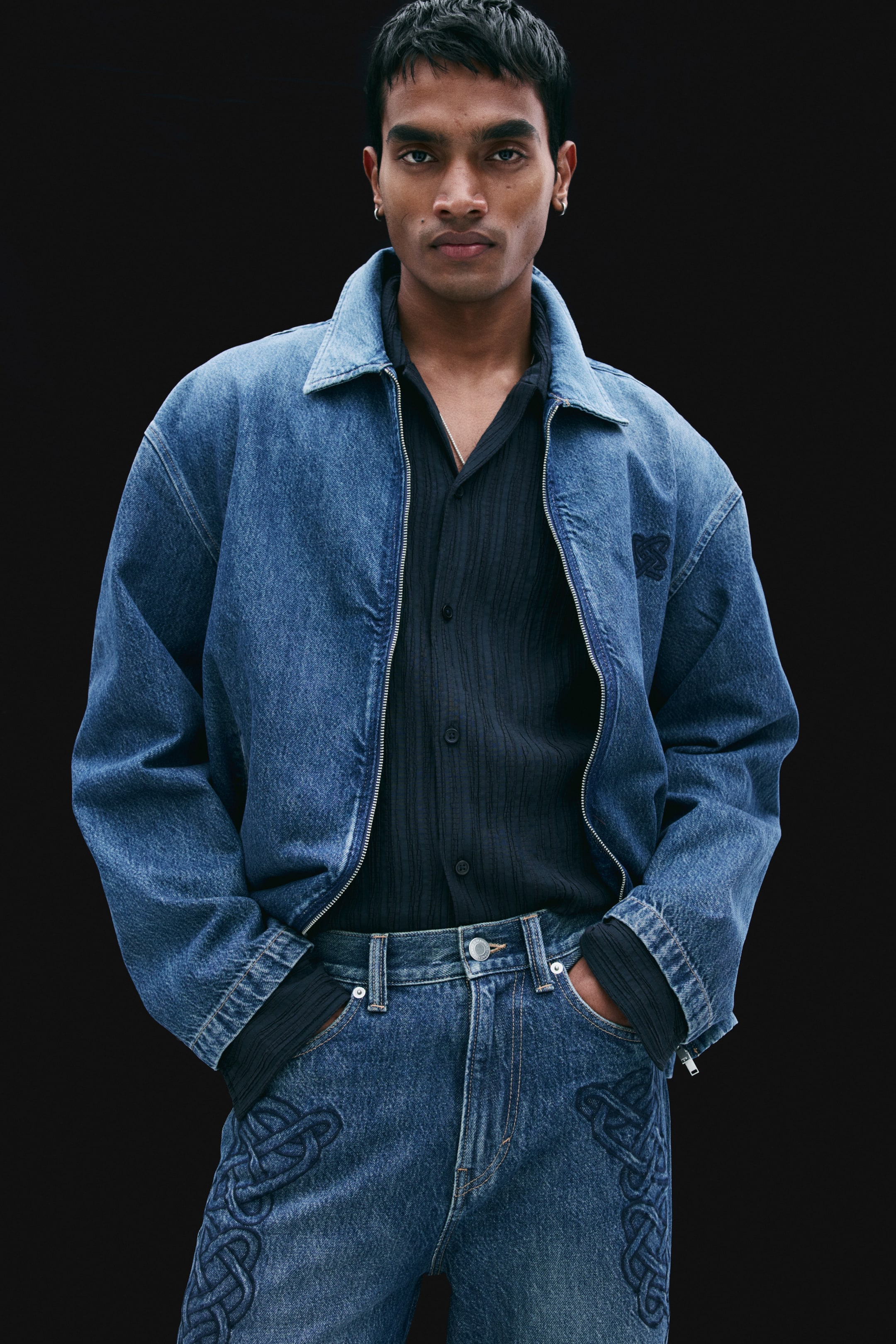View larger image: Blue denim jeans featuring dark-toned embroidered knot patterns on the upper thighs are being worn by the model, who is resting their hands inside the front pockets. The jeans are styled with a tucked-in black ribbed shirt and an open matching blue denim jacket.
