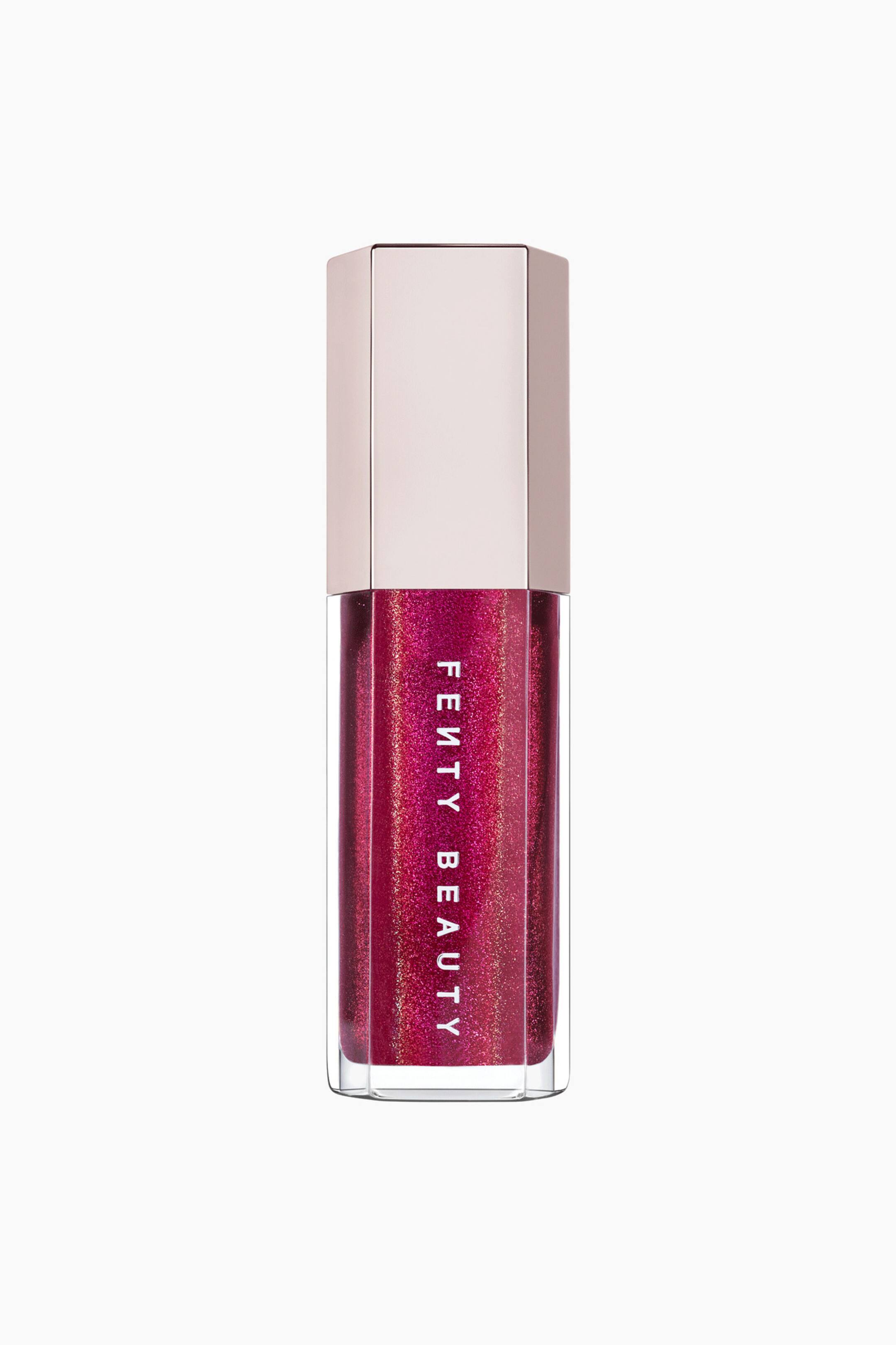 Näytä suurempi kuva: Fenty Beauty -huulikiillon tuubi seisoo pystyssä ja esittelee kirkkaan kuusikulmaisen säiliön, joka on täynnä säihkyvää, syvän magentansävyistä huulikiiltoa. Säiliössä on brändin nimi "FENTY BEAUTY" painettuna pystysuoraan valkoisilla suuraakkosilla, ja sitä viimeistelee metallinhohtoinen, vaalean ruusukullan sävyinen kuusikulmainen korkki.