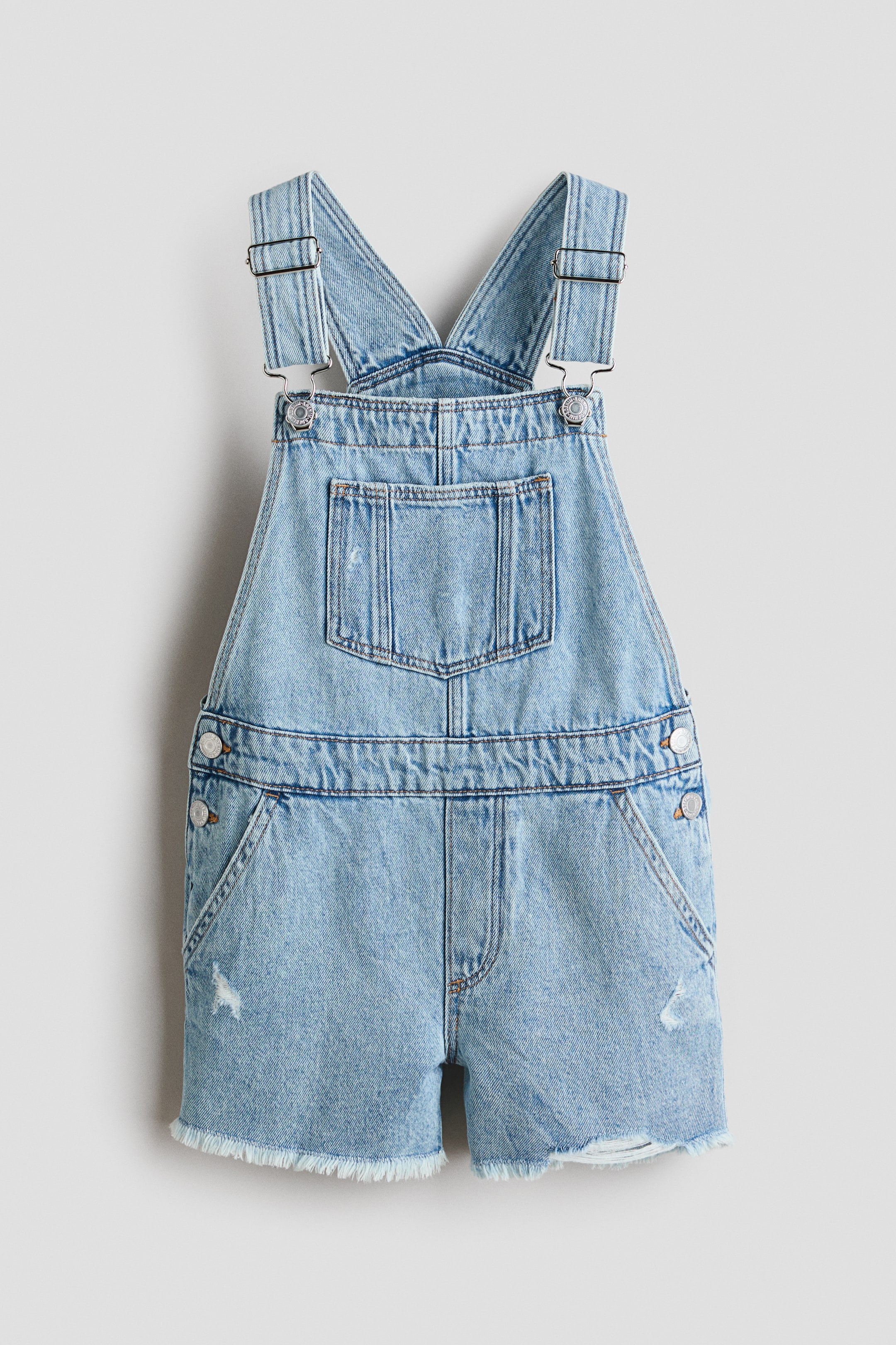 Grösseres Bild ansehen: Hellblaue Denim-Short-Overalls mit Frontansicht, ausgestattet mit verstellbaren Schulterträgern mit Metallverschlüssen, einer Latz-Brusttasche, seitlichen Knopfverschlüssen und abgenutzten Details an den Shorts mit ausgefranstem Saum.