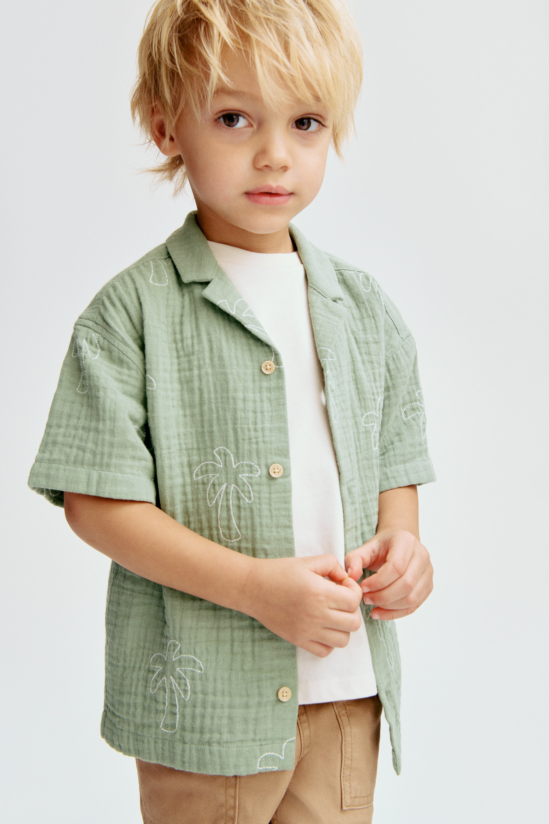 View larger image: A sage green short-sleeved shirt features a textured fabric with white embroidered palm trees and wave-like shapes. It boasts an open camp collar and light-colored buttons down the front, styled open over a white t-shirt and brown pants.