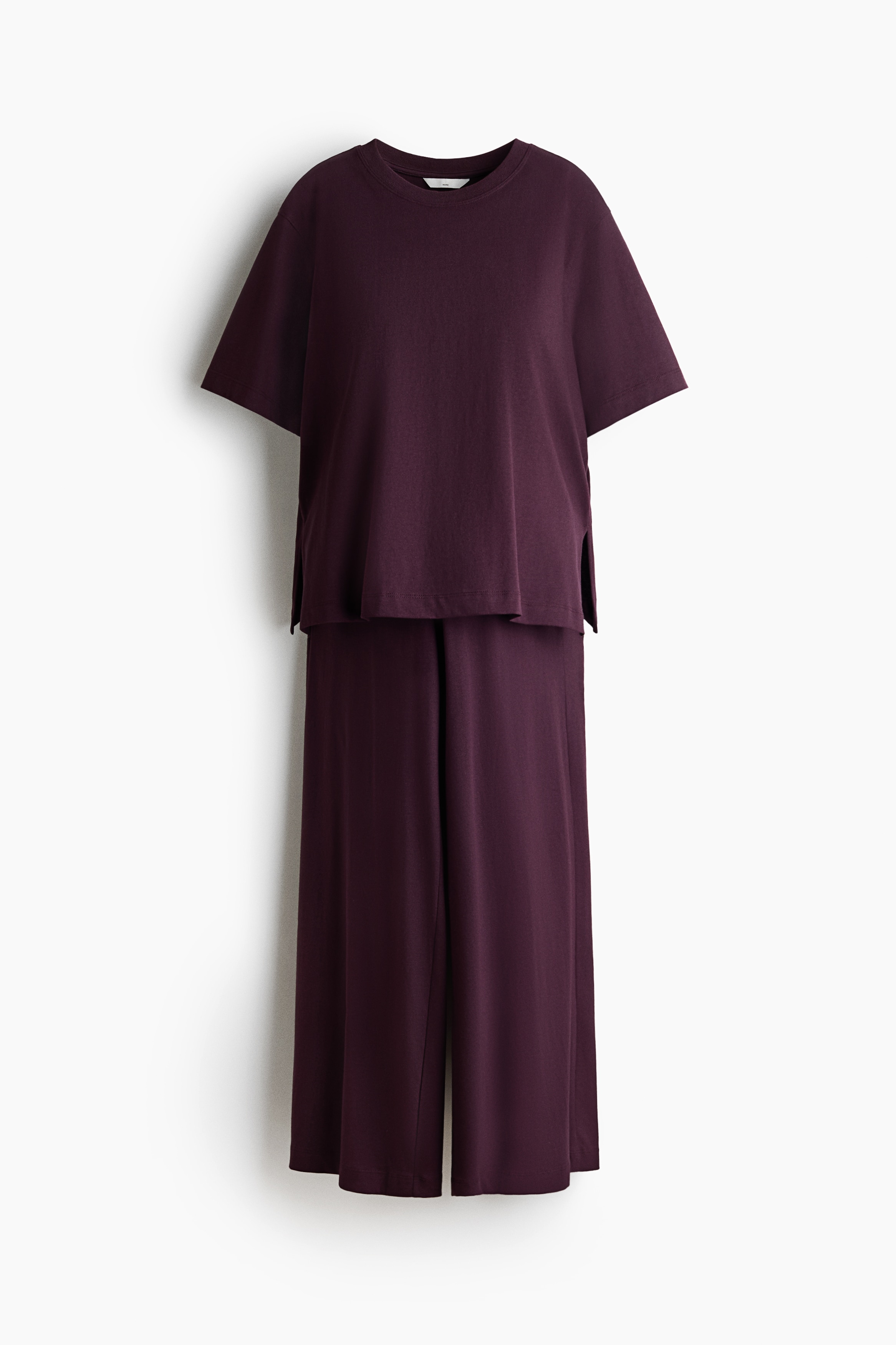 The model is wearing a dark purple T-shirt and trousers set while holding a baby. The relaxed-fit T-shirt features dropped shoulders and a side-split hem, paired with matching wide-leg trousers.