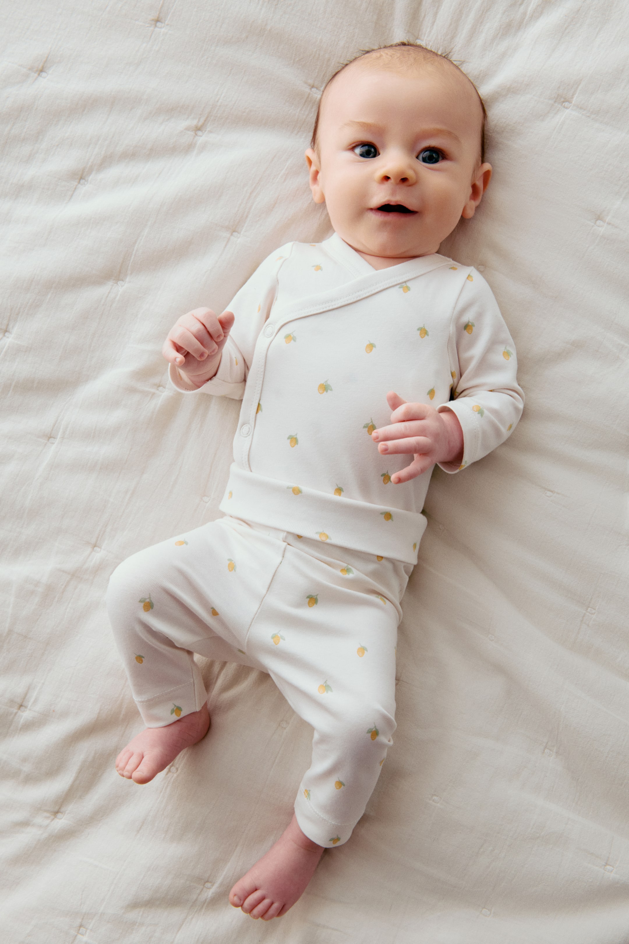 이미지 확대 보기: 크림색 아기 의상이 소개되고 있습니다. 이 의상은 랩 스타일의 긴 소매 탑과 매칭되는 바지로 구성되어 있습니다. 이 의상은 작은 노란색 레몬 모양의 과일과 초록색 잎이 반복되는 패턴을 보여주고 있으며, 탑은 옆면에 스냅으로 잠글 수 있고 바지는 넓은 허리 밴드를 가지고 있습니다.