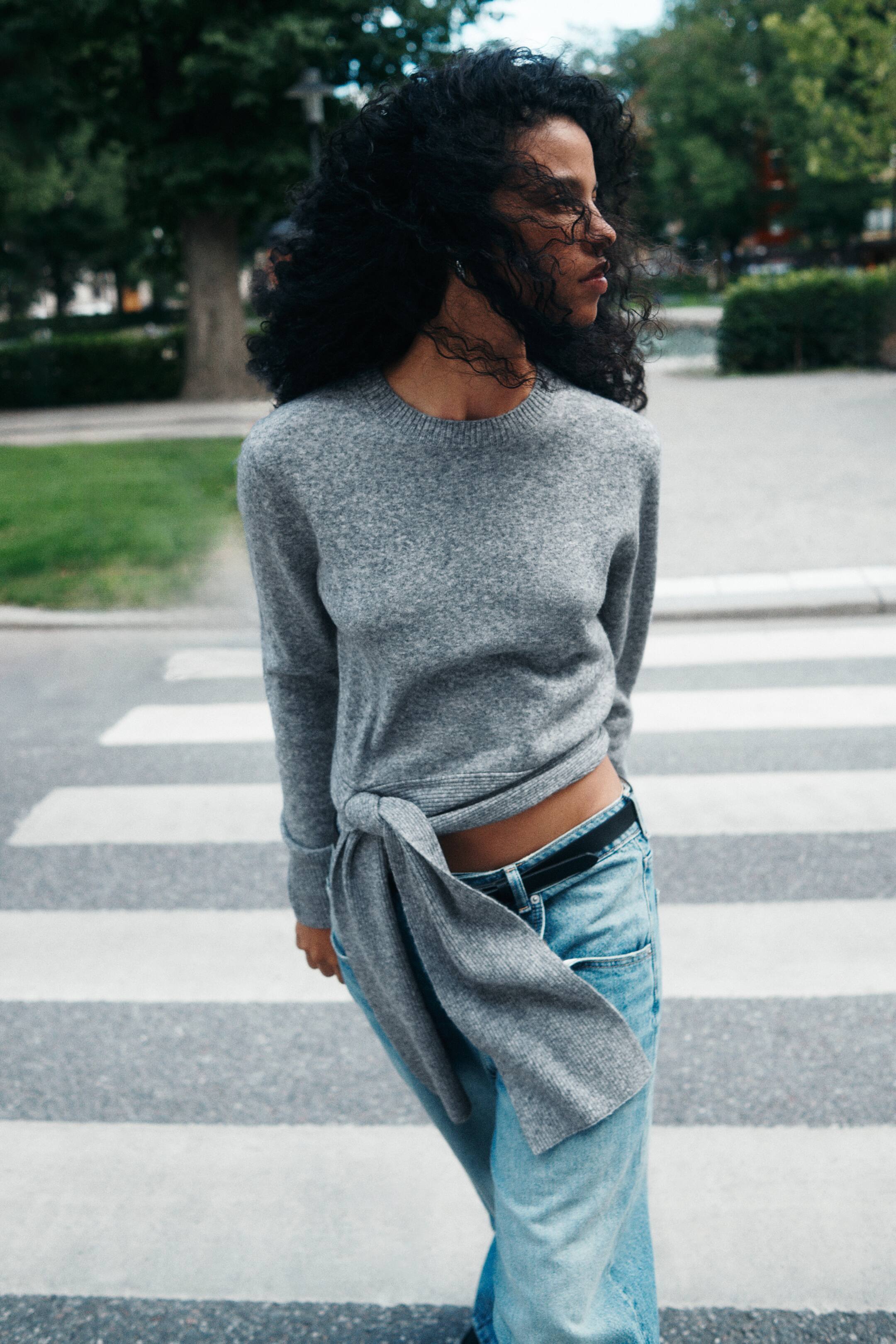 Ver imagen más grande: La modelo lleva un jersey de punto fino gris, con cuello redondo acanalado, mangas largas con puños acanalados y un bajo con lazadas anchas anudadas en el lateral delantero. El jersey se combina con unos vaqueros azul claro y un cinturón negro, dejando ver parte de la cintura.