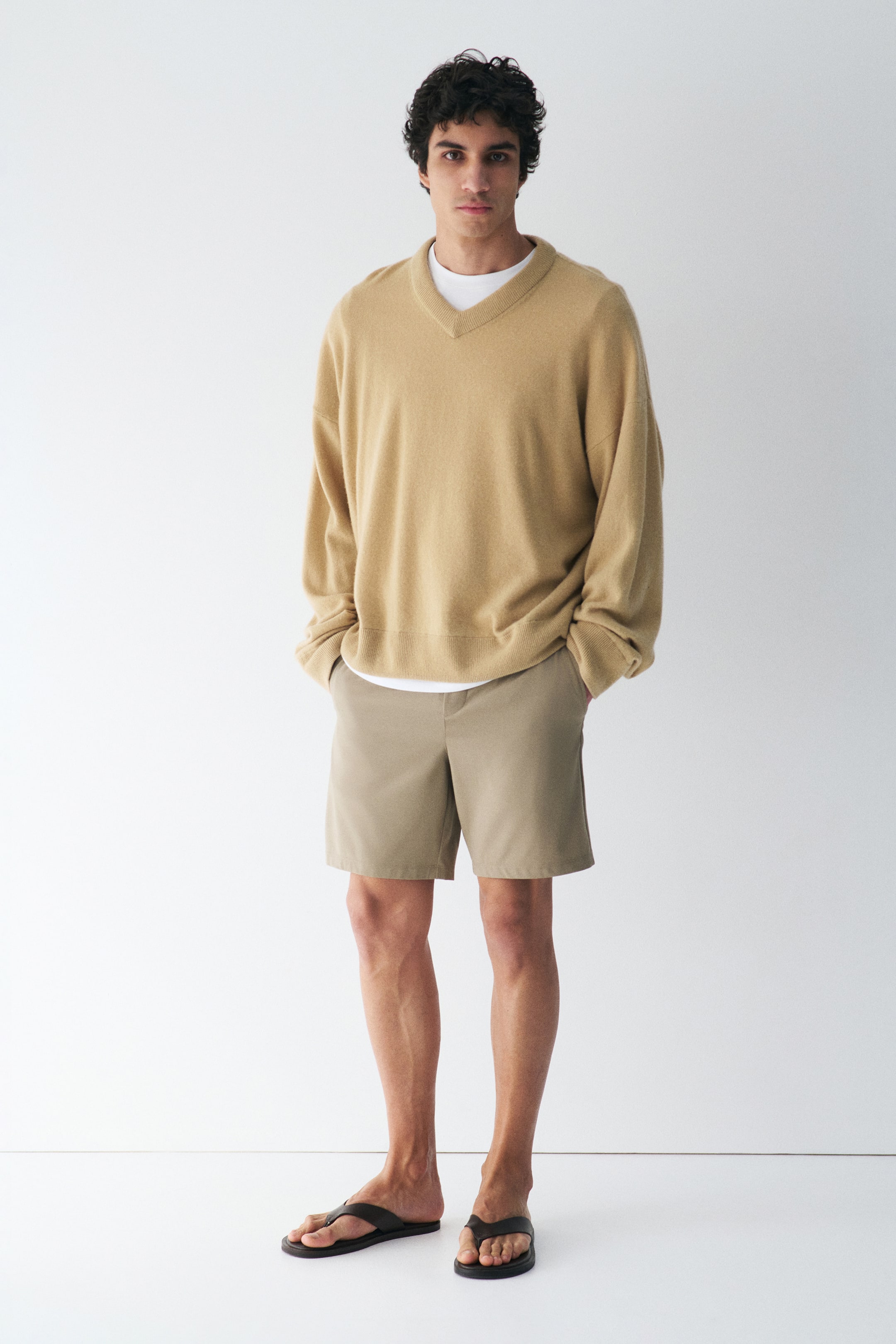 View larger image: A beige V-neck knit sweater is worn over a white t-shirt, showcasing a relaxed, oversized fit with long sleeves and a straight hem. Beige flat-front shorts are worn, extending above the knee, and dark flip-flops complete the look.