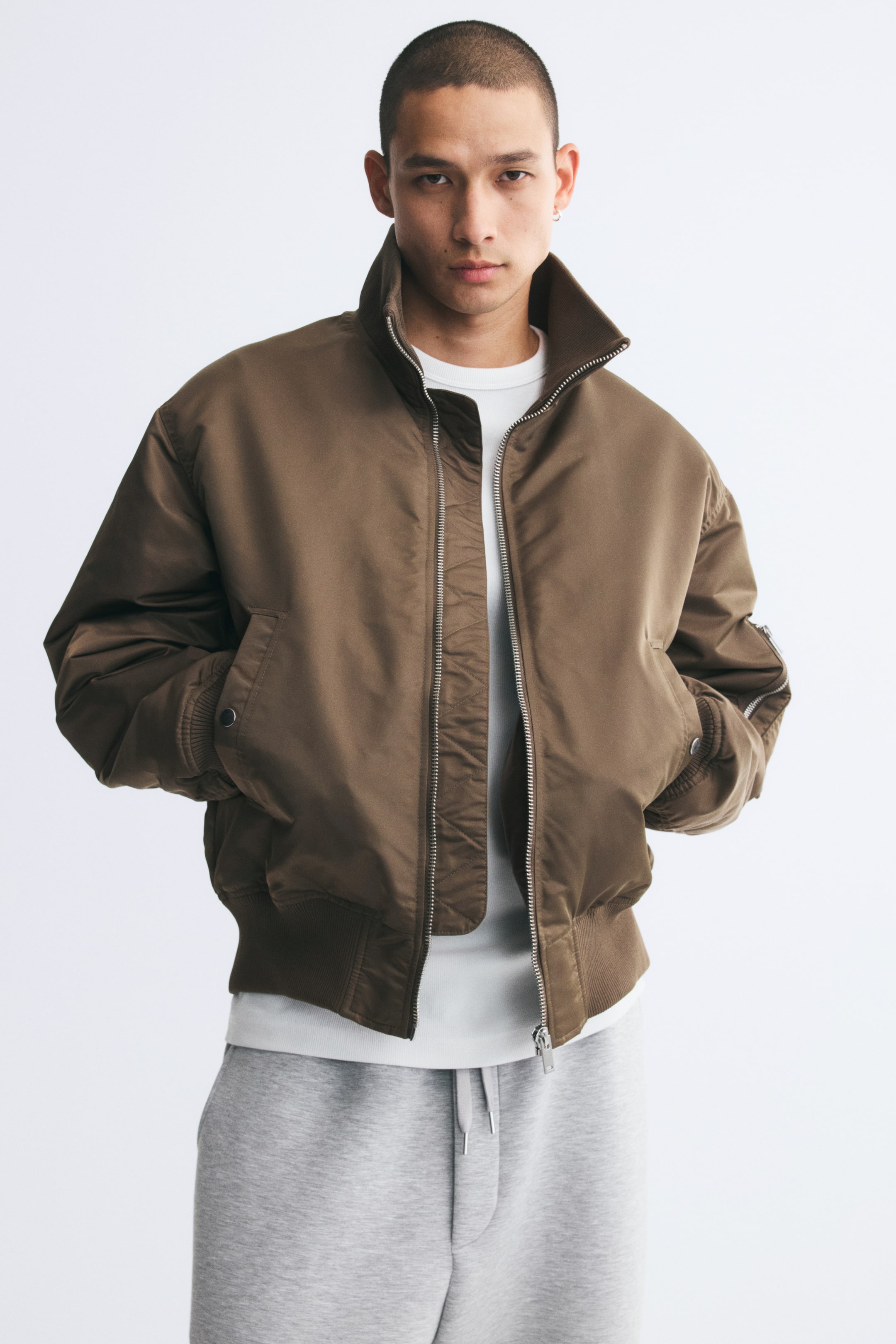 Visualizza immagine più grande: Una giacca bomber in nylon imbottito color marrone è indossata dalla modella, che tiene le mani nelle tasche a filetto anteriori. La giacca presenta una zip frontale aperta, colletto alto a costine, spalle scese e una tasca sulla manica con zip, ed è abbinata a una T-shirt bianca e a pantaloni della tuta grigi.