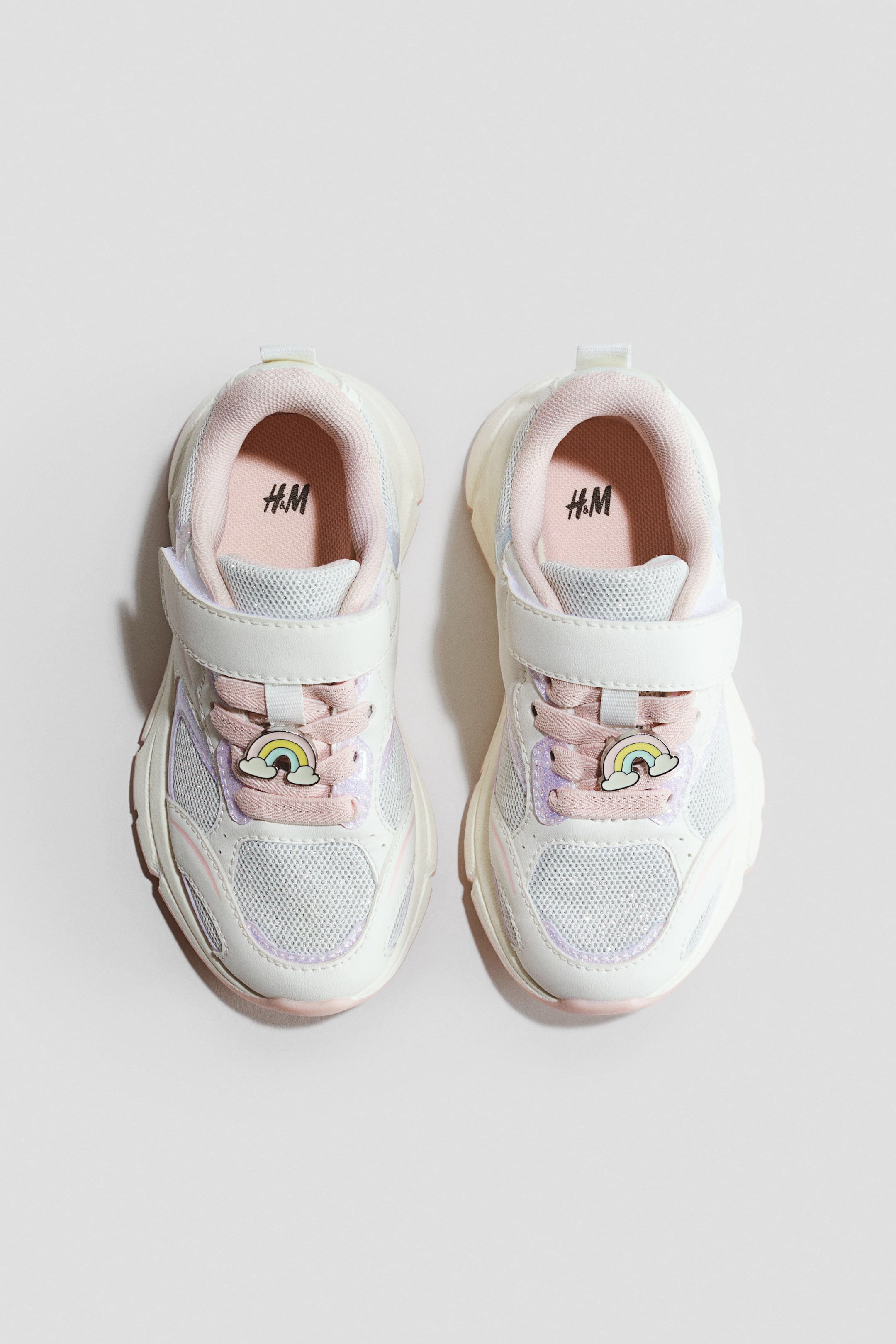 Ingrandisci l'immagine: Un paio di sneakers per bambini è mostrato frontalmente, caratterizzato da pannelli in mesh bianco e sovrapposizioni sintetiche. Ogni scarpa presenta lacci rosa fissati da una chiusura a strappo bianca e un piccolo charm arcobaleno. Le solette visibili sono rosa chiaro e includono il logo H&M.