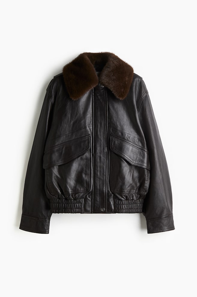 Une veste bomber en cuir marron foncé présente son devant, avec un col rabattu en fourrure marron foncé proéminent. Elle met en avant deux grandes poches à rabat sur le bas du devant et un ourlet élastiqué froncé. - 1