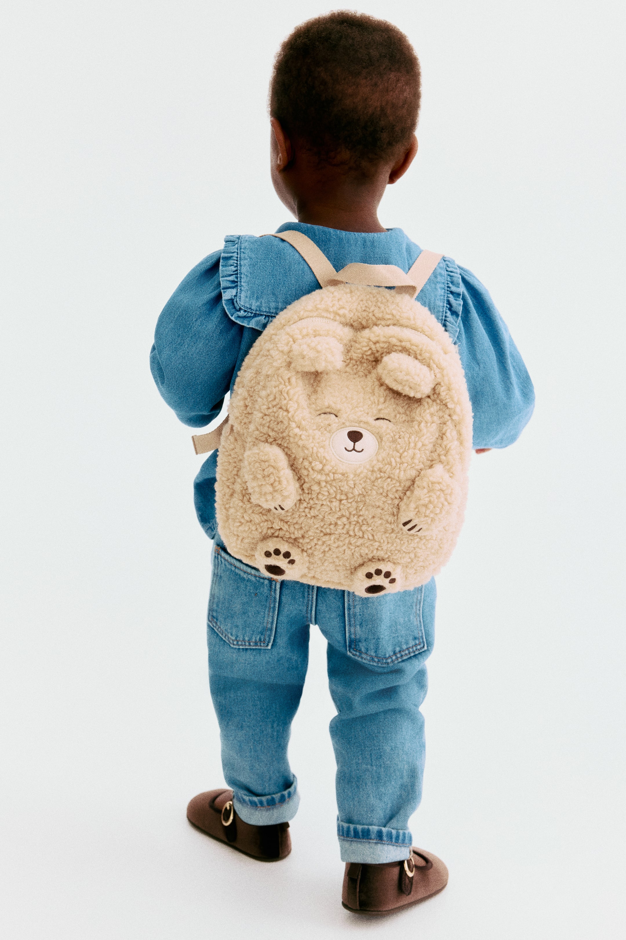 Visa större bild: En mjuk teddyryggsäck visas bakifrån, designad med ett lekfullt björnansikte med broderade detaljer och tassar. Den är utrustad med ett handtag upptill, dragkedja och justerbara axelremmar i webbing.