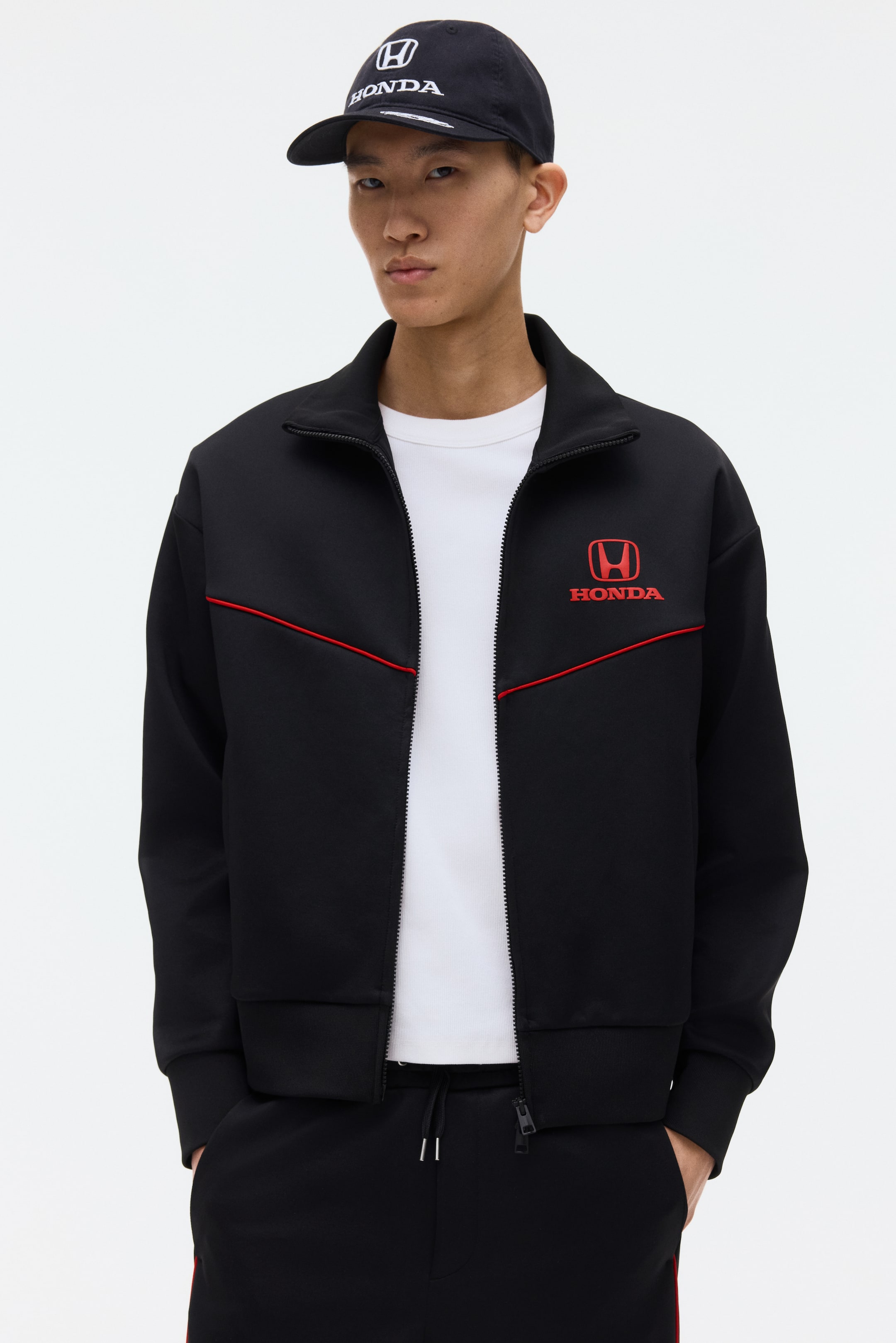 View larger image: A black track jacket showcasing a red Honda logo and 'HONDA' text on the left chest, complemented by a red stripe running horizontally across the front. It features a full-zip closure, a stand-up collar, ribbed cuffs, and a ribbed hem, with a white t-shirt peeking underneath.