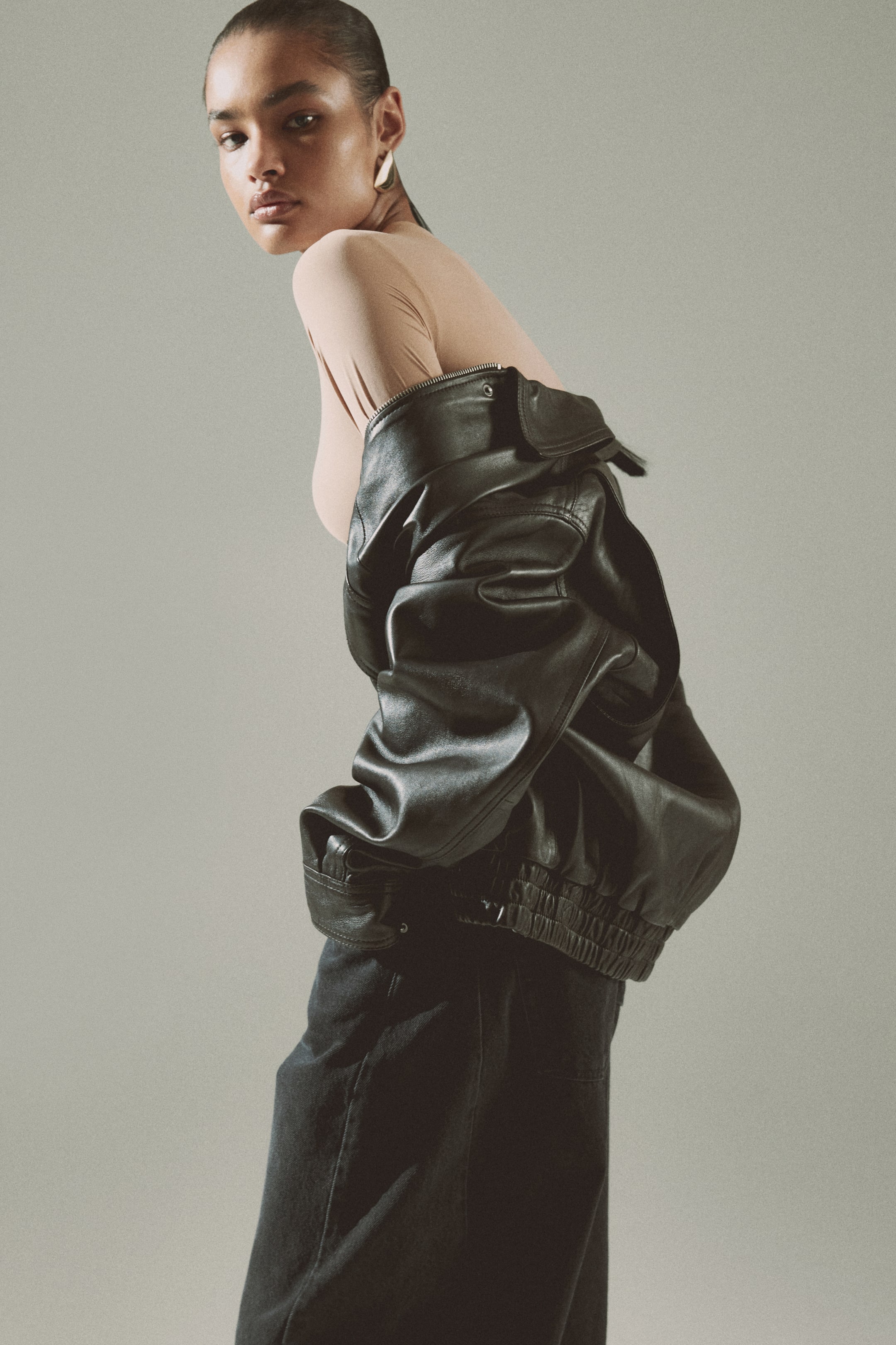 View larger image: A black leather bomber jacket is draped over the left shoulder, highlighting its side-back view. It features a prominent silver-toned zip closure at the front and an elasticised hem, layered over a light-coloured top and dark trousers.