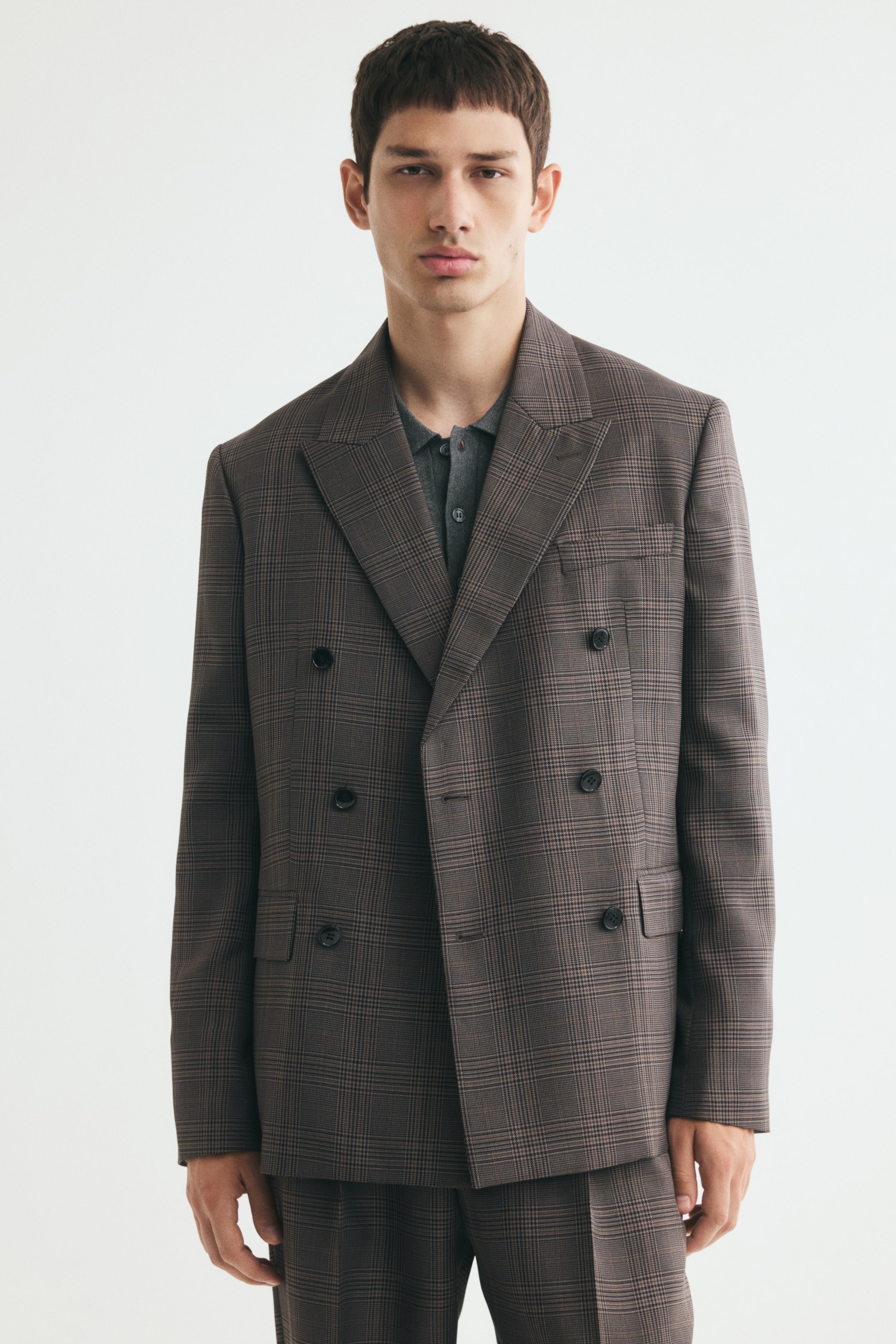 View larger image: The model is wearing a brown checked double-breasted jacket over a grey collared shirt and matching trousers. The jacket features broad peak lapels, a chest pocket, front flap pockets, and a six-button front fastening.